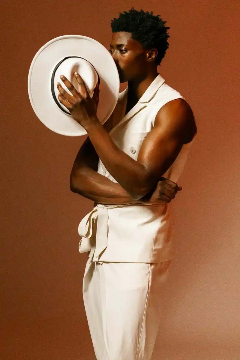 Artistic portrait of a person in white outfit holding a wide-brimmed hat against a brown backdrop.