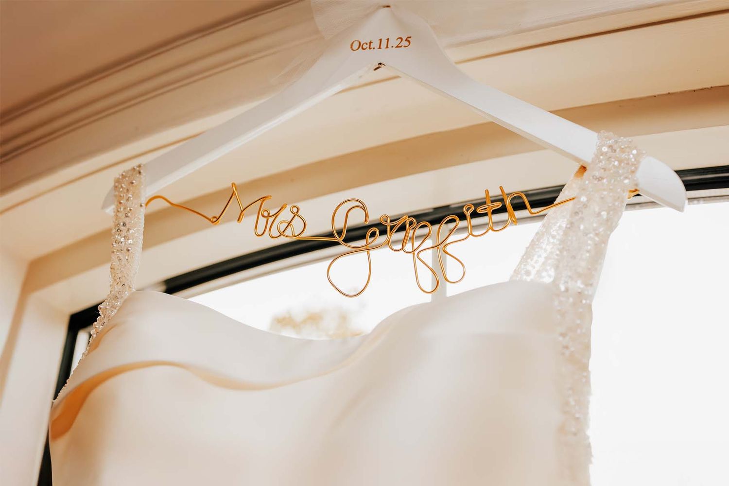 A white wedding dress hanging on a personalized hanger in natural light.