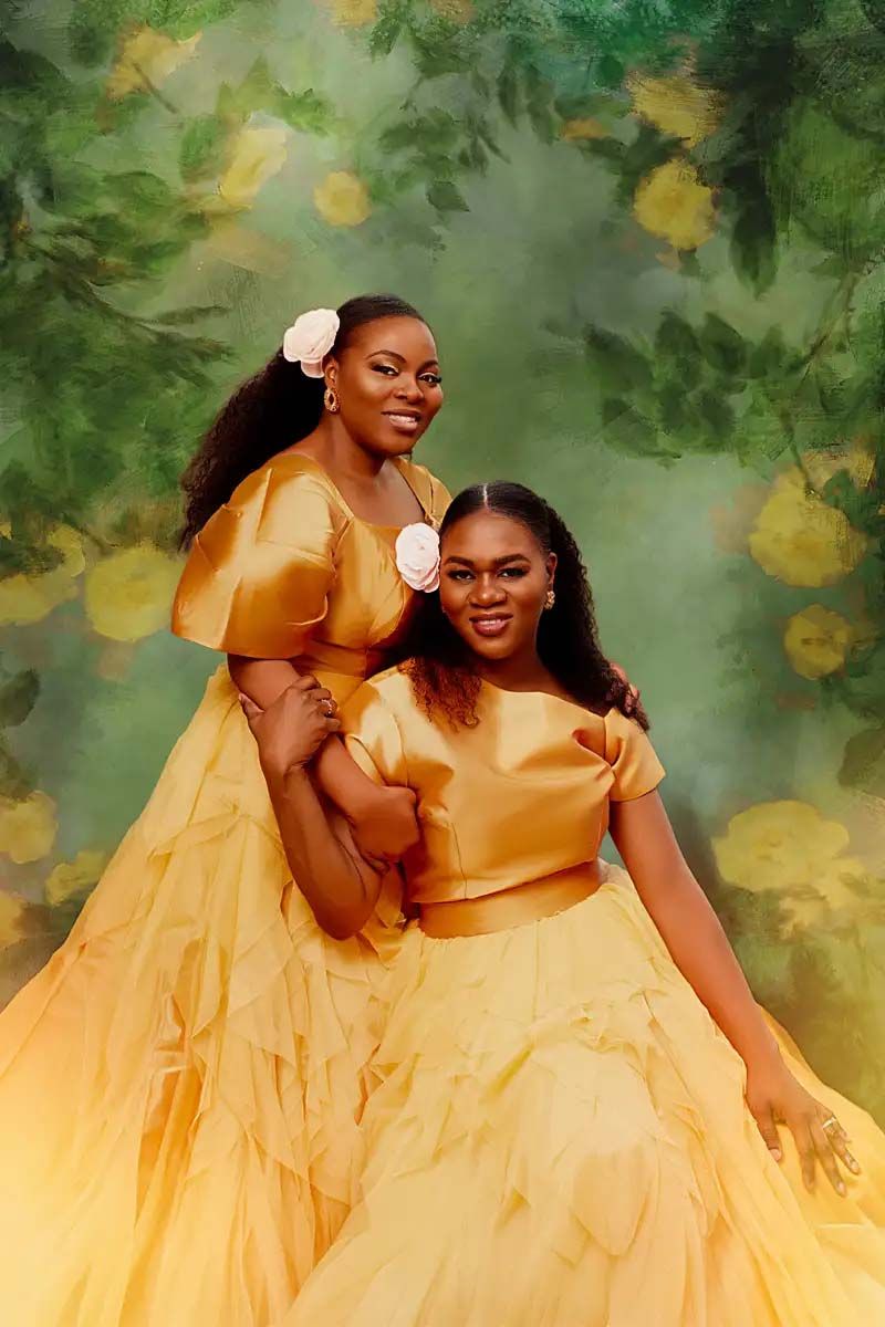 Two models in flowing yellow gowns pose together against a dreamy garden backdrop.