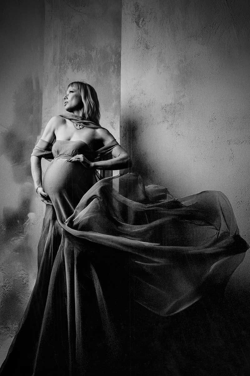 Artistic black and white maternity portrait with flowing fabric against textured wall.