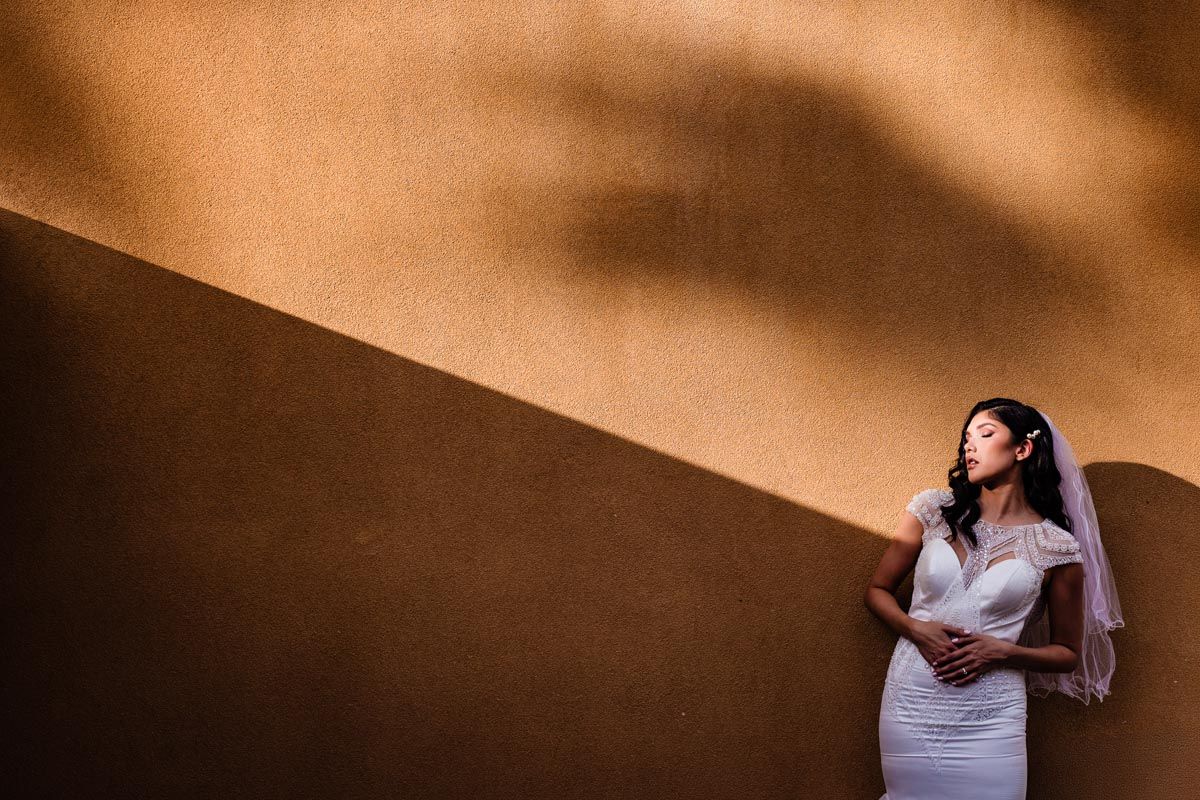 A bride in white dress poses against a textured wall with dramatic angular shadows.