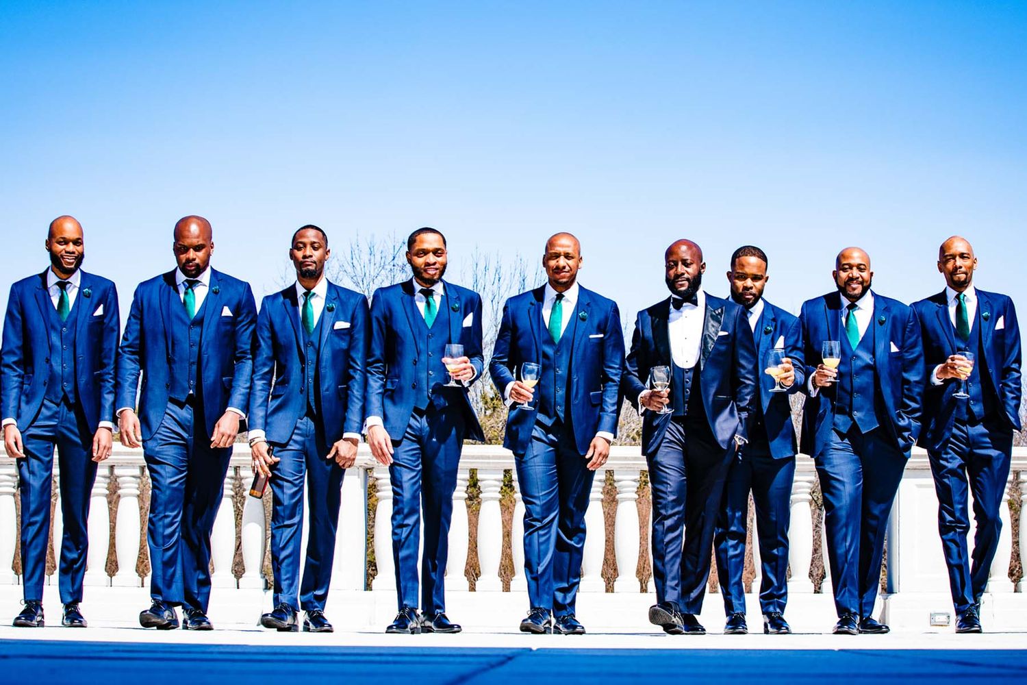 Wedding party in royal blue suits and white dresses posed in a line against a bright blue sky.