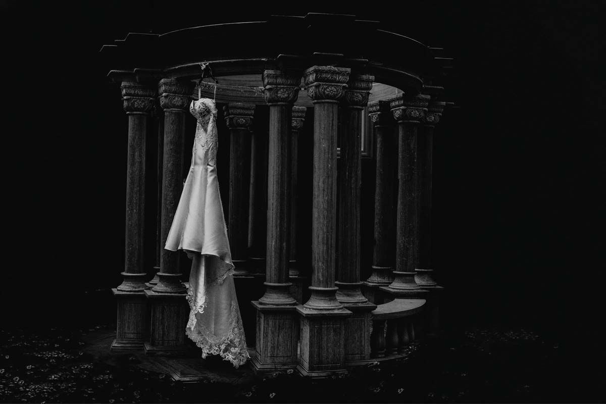 A wedding dress hangs elegantly among dramatic black stone pillars in artistic black and white photography.