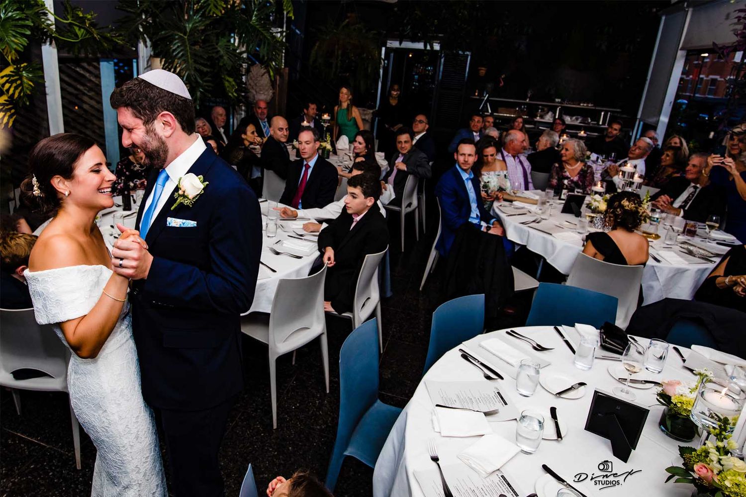 Wedding reception dinner scene with guests seated at decorated tables in dimly lit venue.