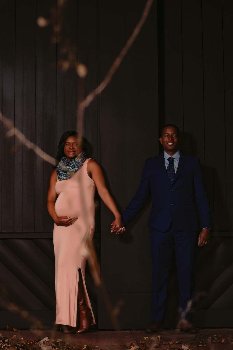 A couple walks together at dusk wearing formal attire against a dark wooden backdrop.