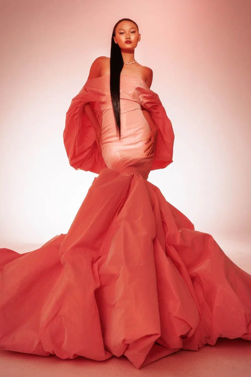 Fashion model in dramatic coral gown with flowing fabric against pink backdrop.