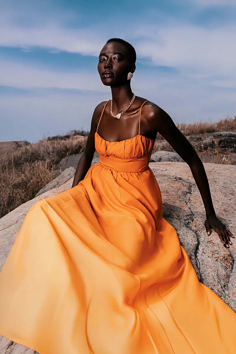 Model in flowing orange dress poses against desert landscape and blue sky.