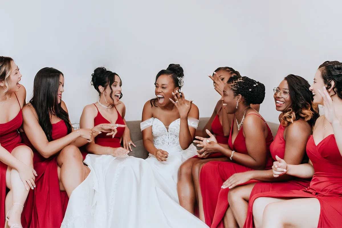 A bridal party in red dresses and white gown share joyful laughter while sitting together.