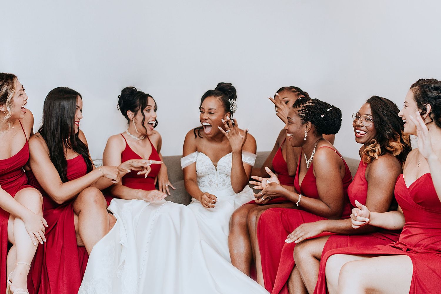 A bride in white dress sits with bridesmaids wearing red dresses sharing laughter and drinks together.