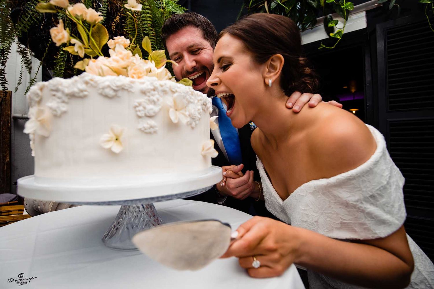 Wedding couple shares a playful moment while cutting their elegant white tiered cake at the reception.