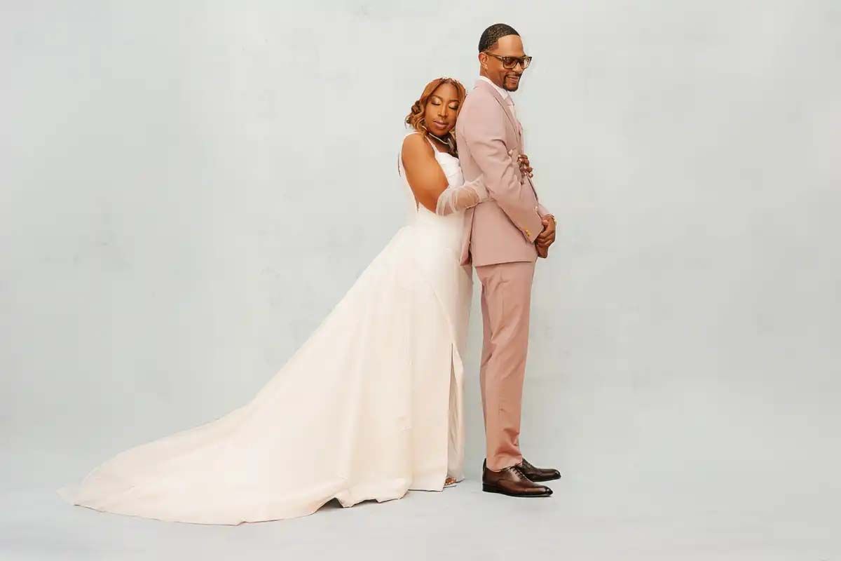 Wedding couple in formal attire poses together on a light neutral background.