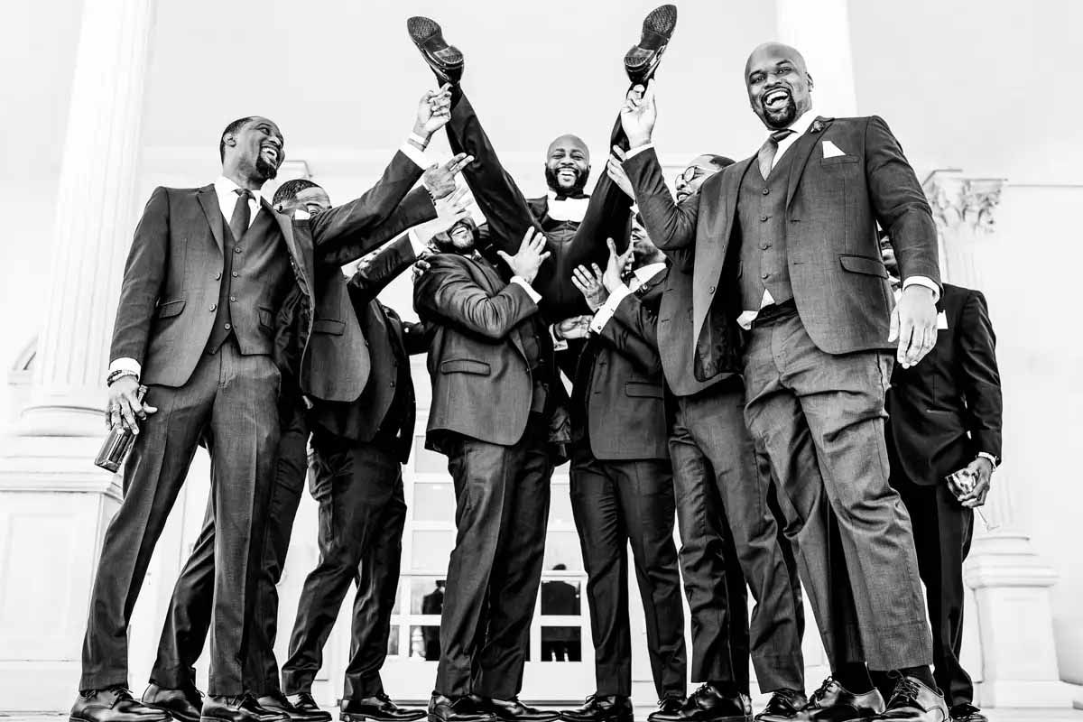 Groomsmen in dark suits celebrate by raising their arms in a joyful black and white photo.
