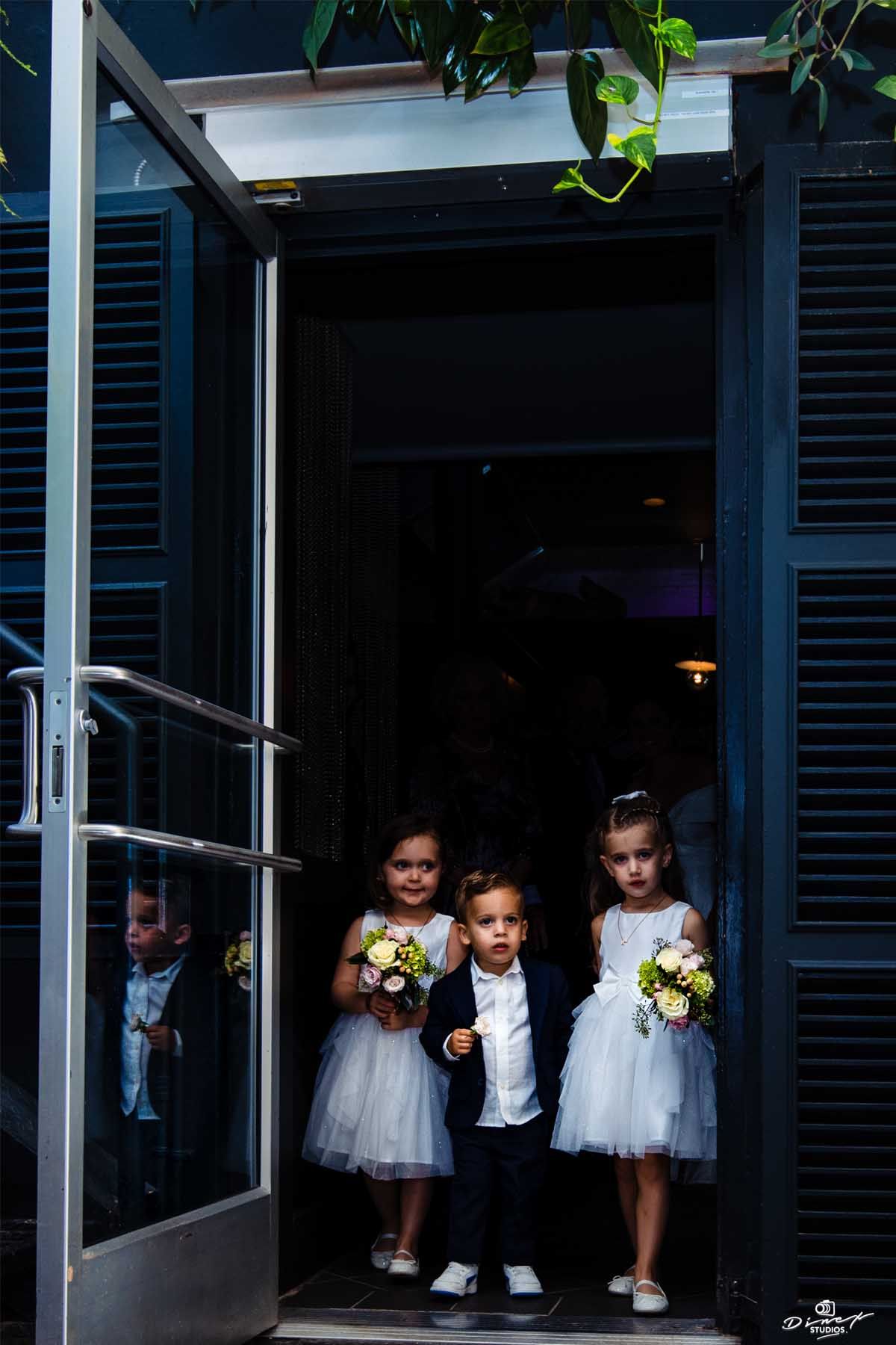 Three young children in formal attire standing in a doorway holding flower bouquets.