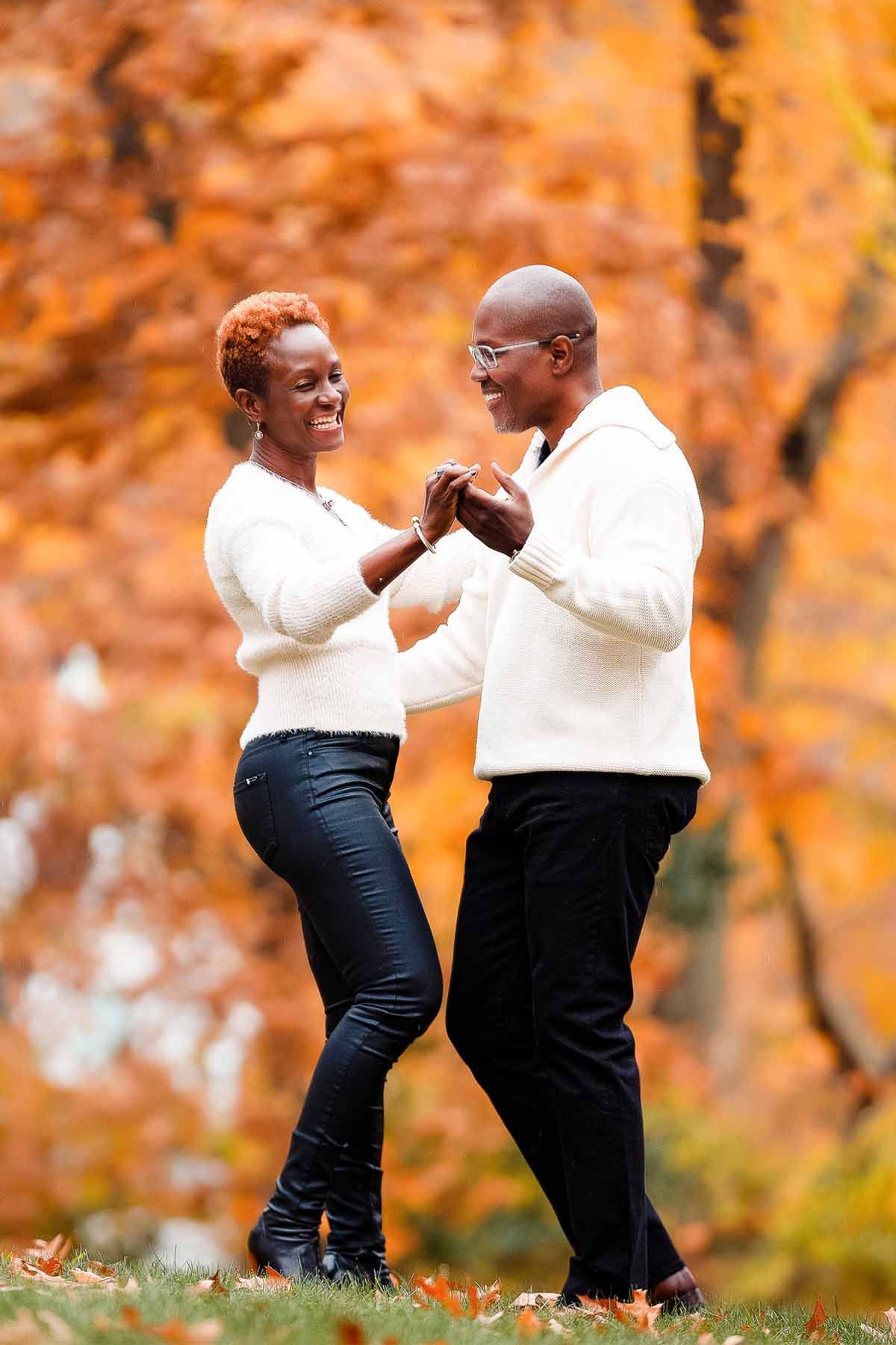 Playful autumn engagement photos with couple in white sweaters against orange foliage.