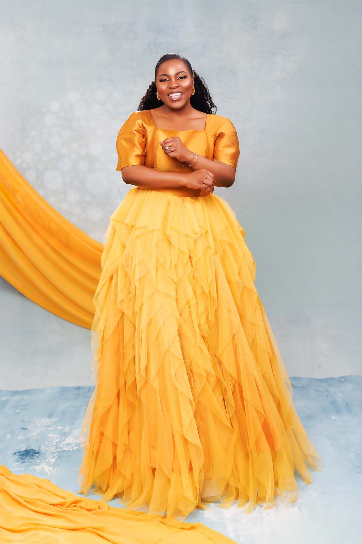 Woman in stunning yellow layered gown poses gracefully against grey background.