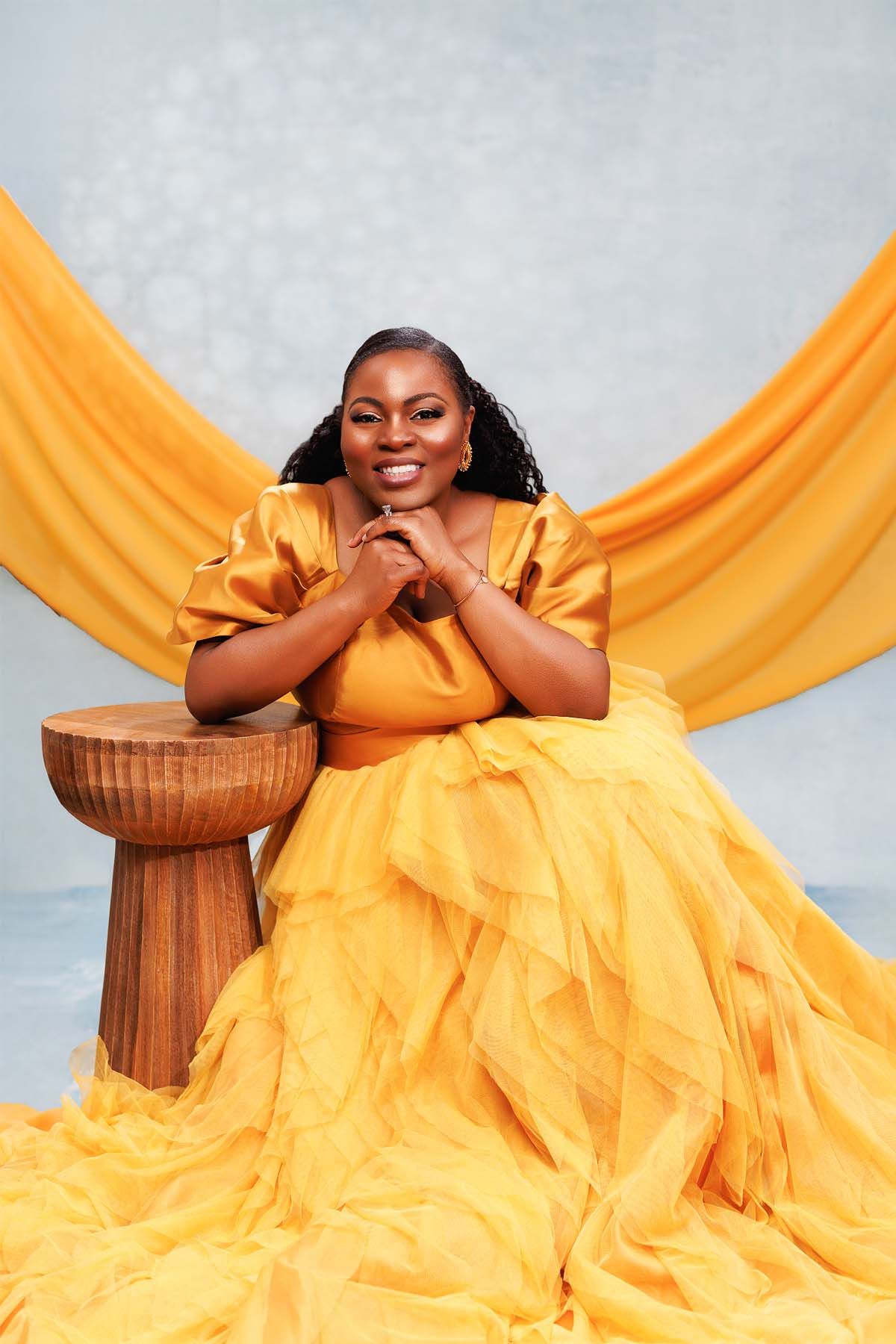 Woman in vibrant yellow gown poses gracefully against draped fabric backdrop.