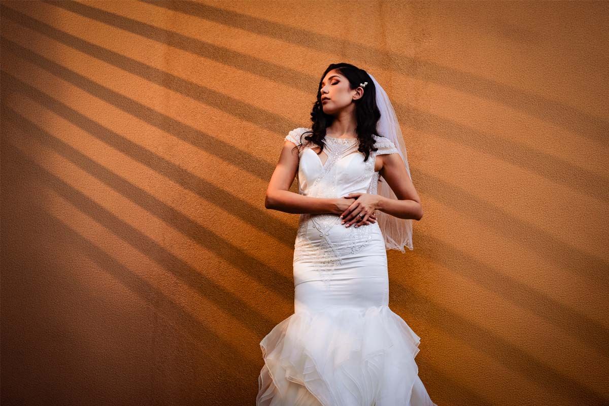 A bride in a fitted mermaid gown poses against a wall with striking diagonal light patterns.