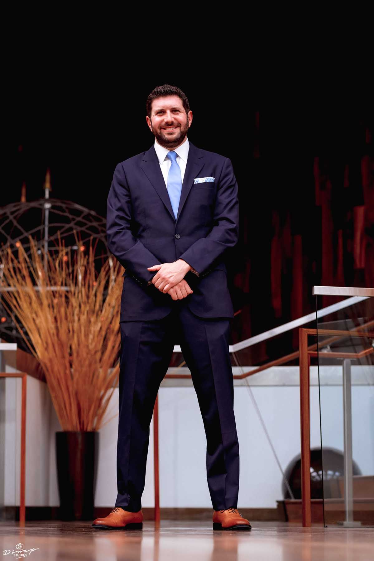 A groom in a navy suit standing confidently on a stage with decorative wheat stalks.