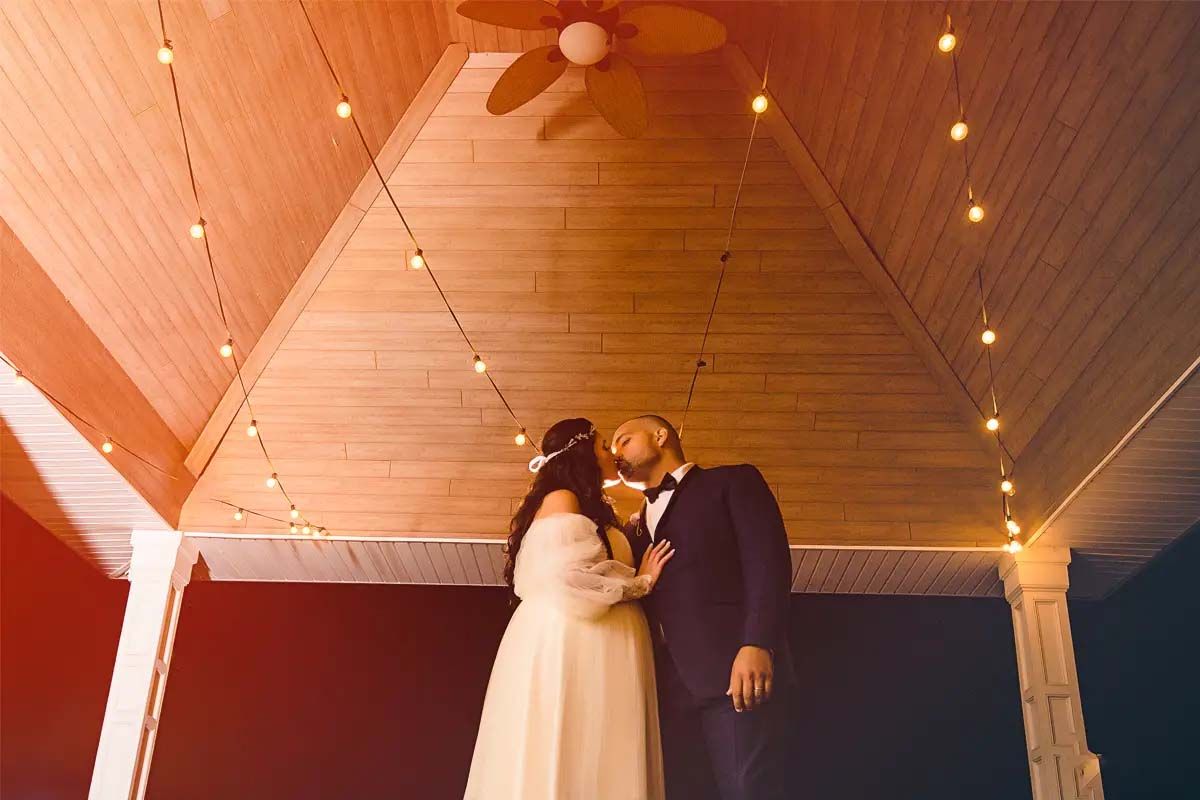 A romantic couple shares an intimate moment under twinkling lights on a covered porch.