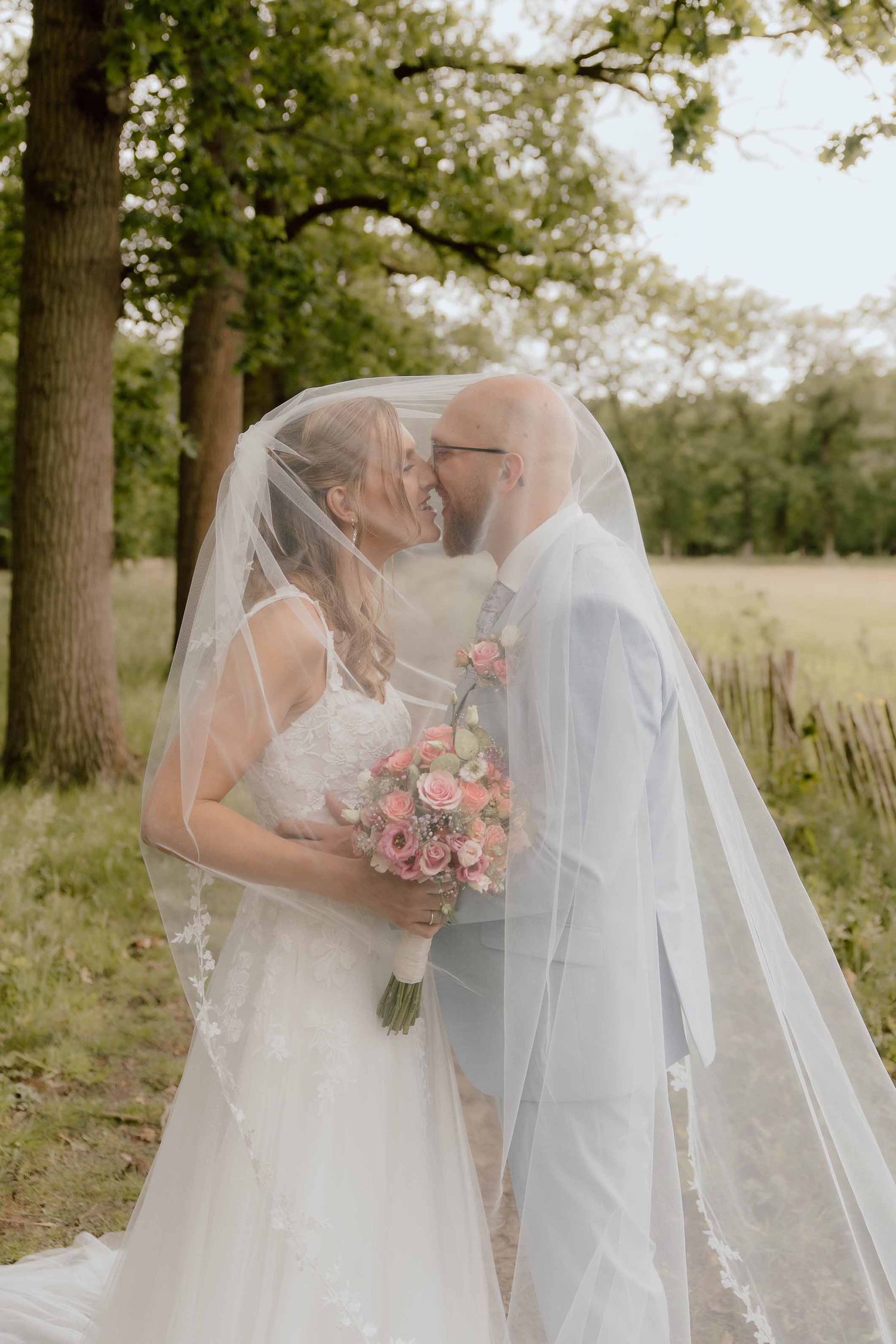 Bruid en bruidegom geven elkaar een kus onder de sluier tijdens de loveshoot in het bos met hoge bomen