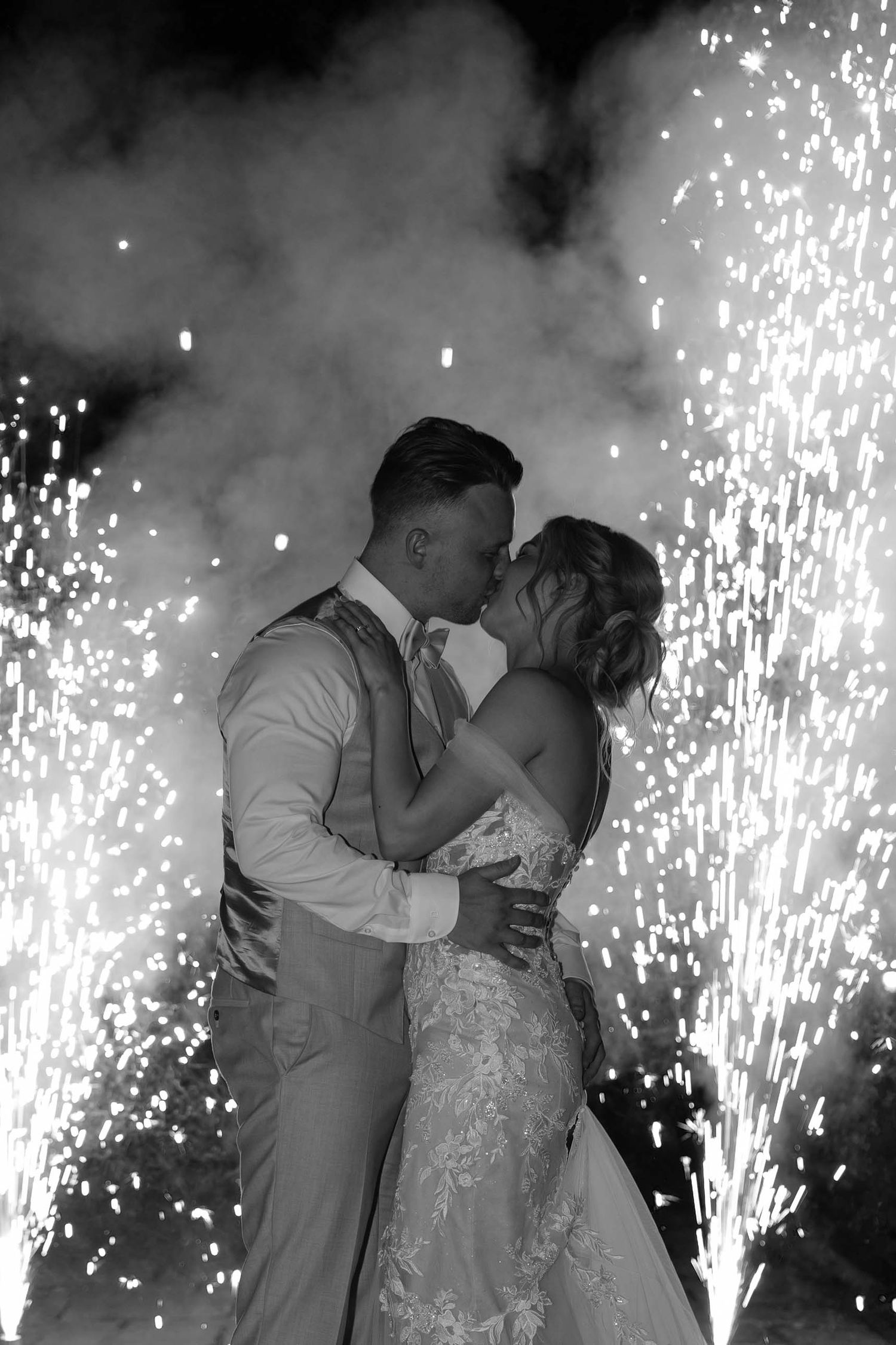 Zwart wit foto van kussend bruidspaar met vuurwerk op de achtergrond