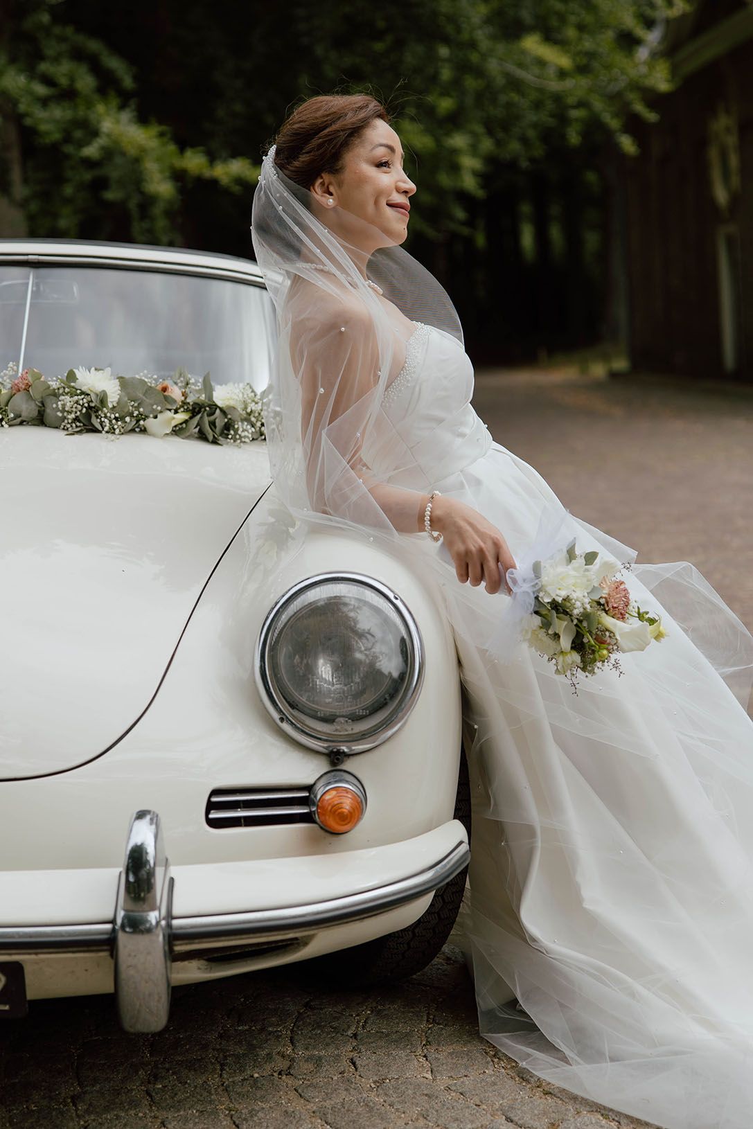 Bruid leunt tijdens de loveshoot tegen de trouwauto met trouwboeket