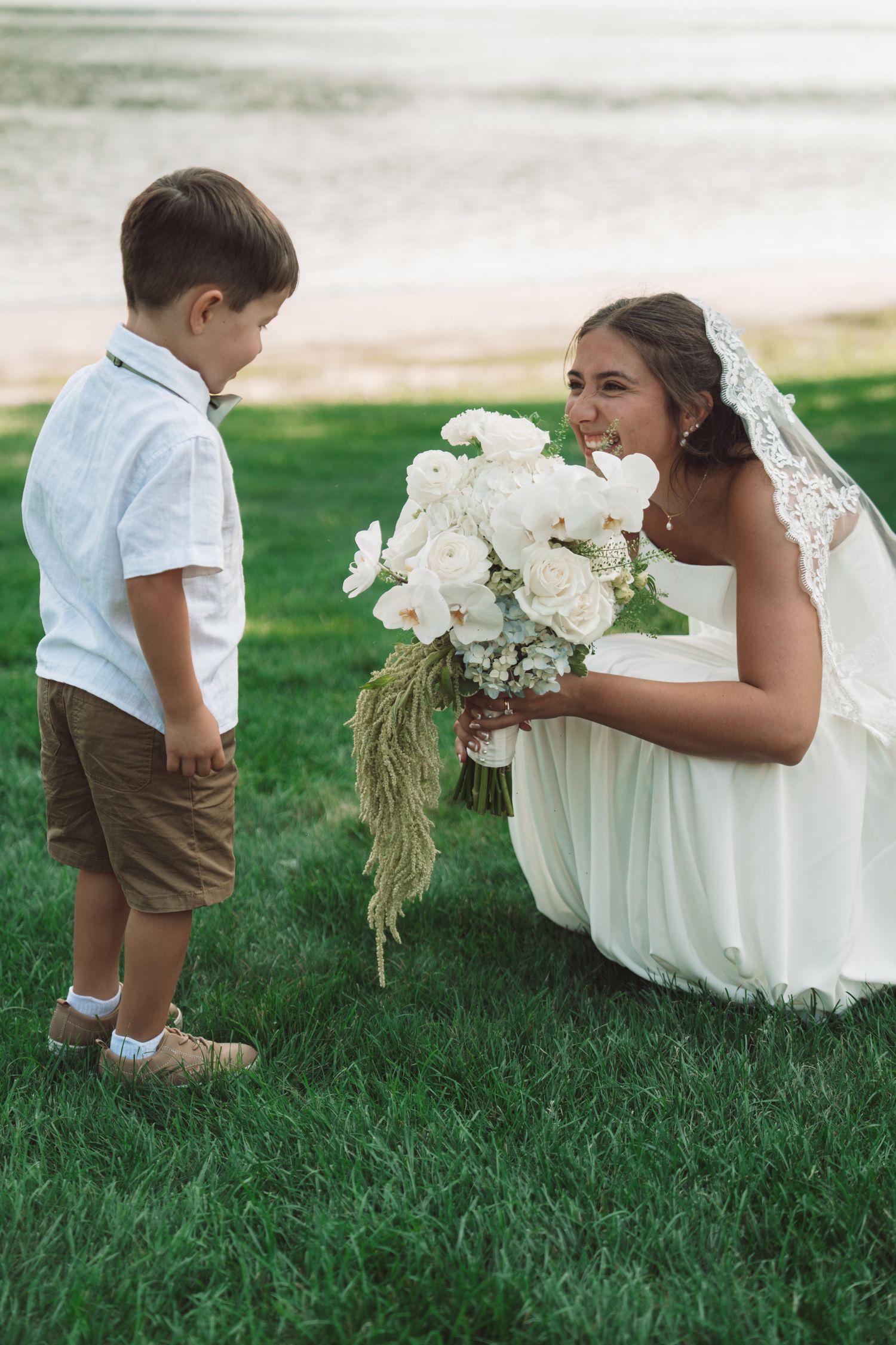 Bride in white gown and veil crouches on green grass, smiling at young boy holding white flower bouquet near waterfront.