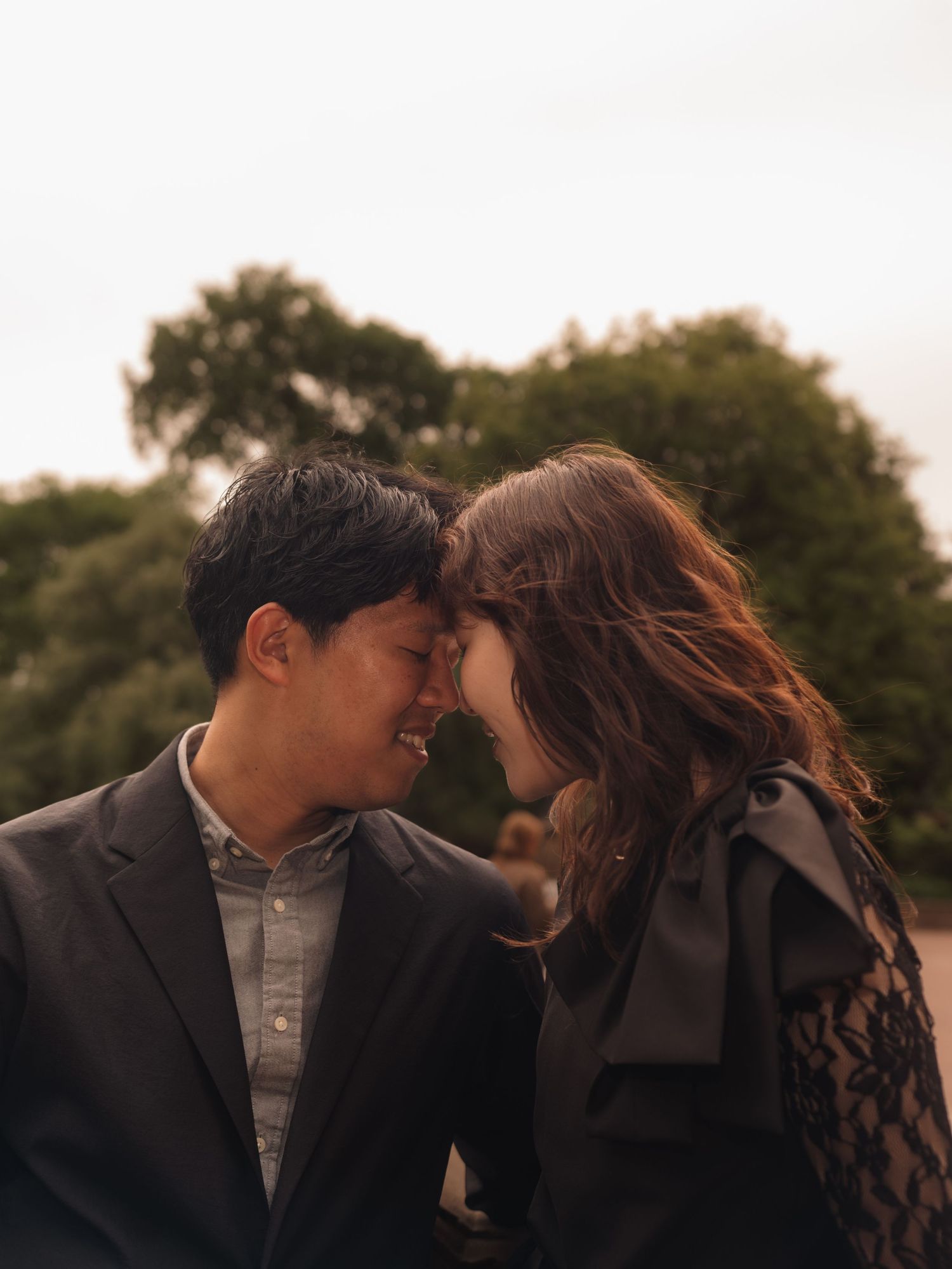 A couple shares a tender forehead-to-forehead moment, smiling softly outdoors with lush green trees in the background.