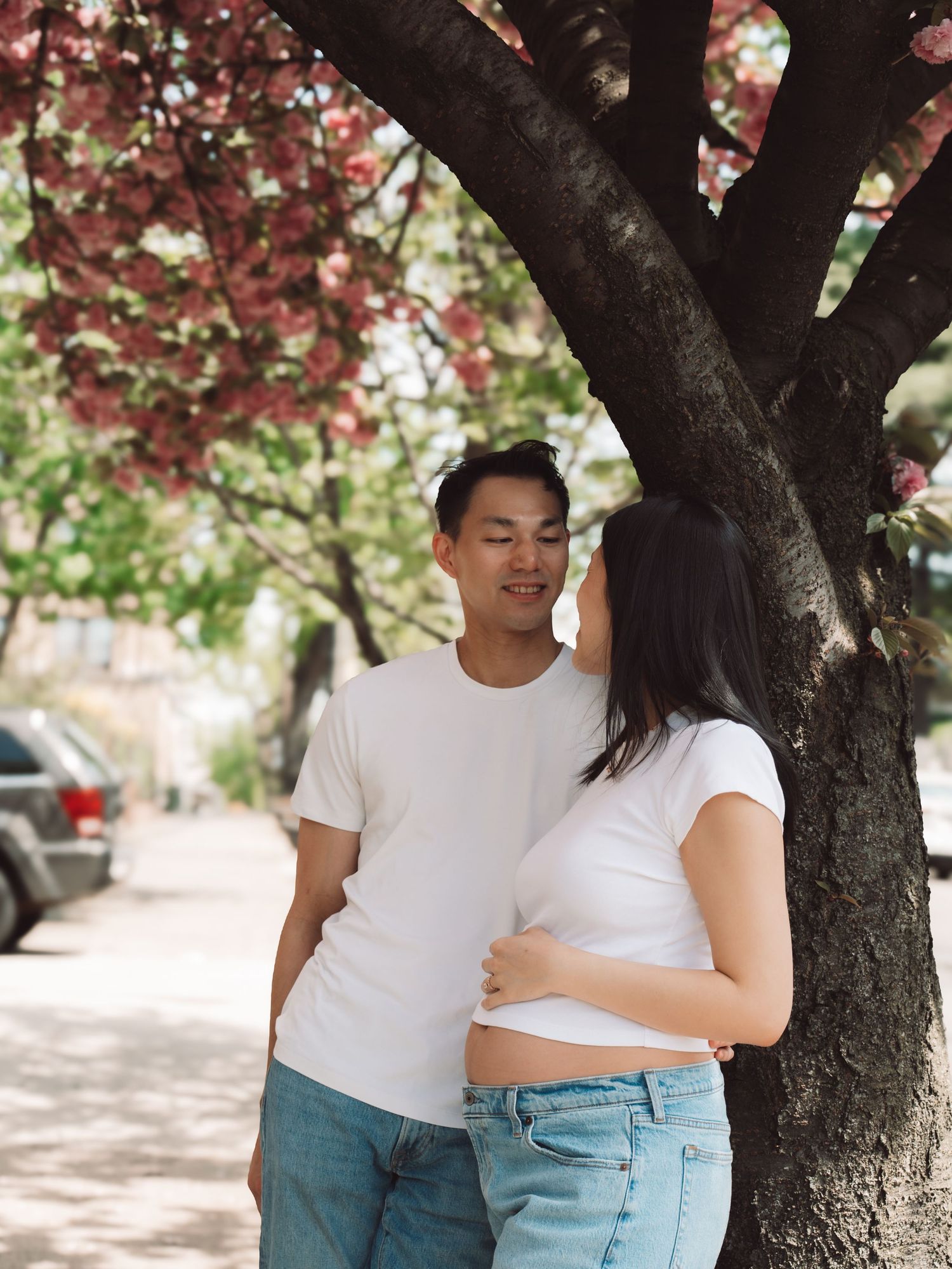 A pregnant couple in matching white tees shares a tender moment under a blooming cherry blossom tree on a sunny street.