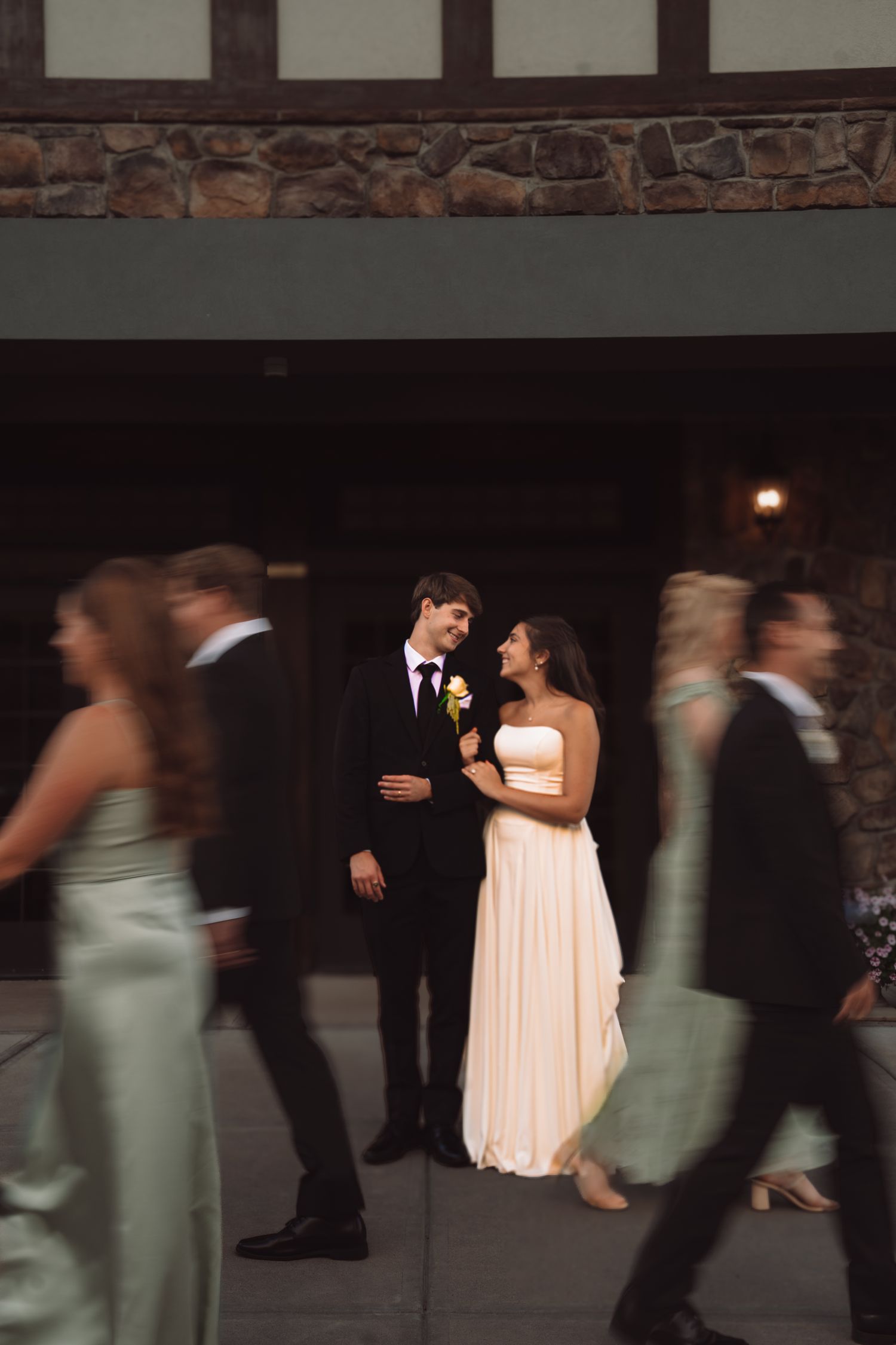 Bride in white gown and groom in black tuxedo share a tender moment as guests walk past in elegant attire.