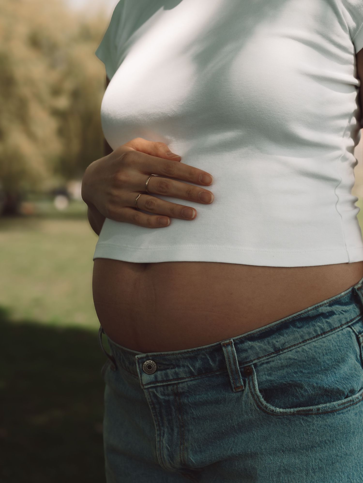 Pregnant woman in white crop top and jeans gently cradling her baby bump outdoors in a park setting.