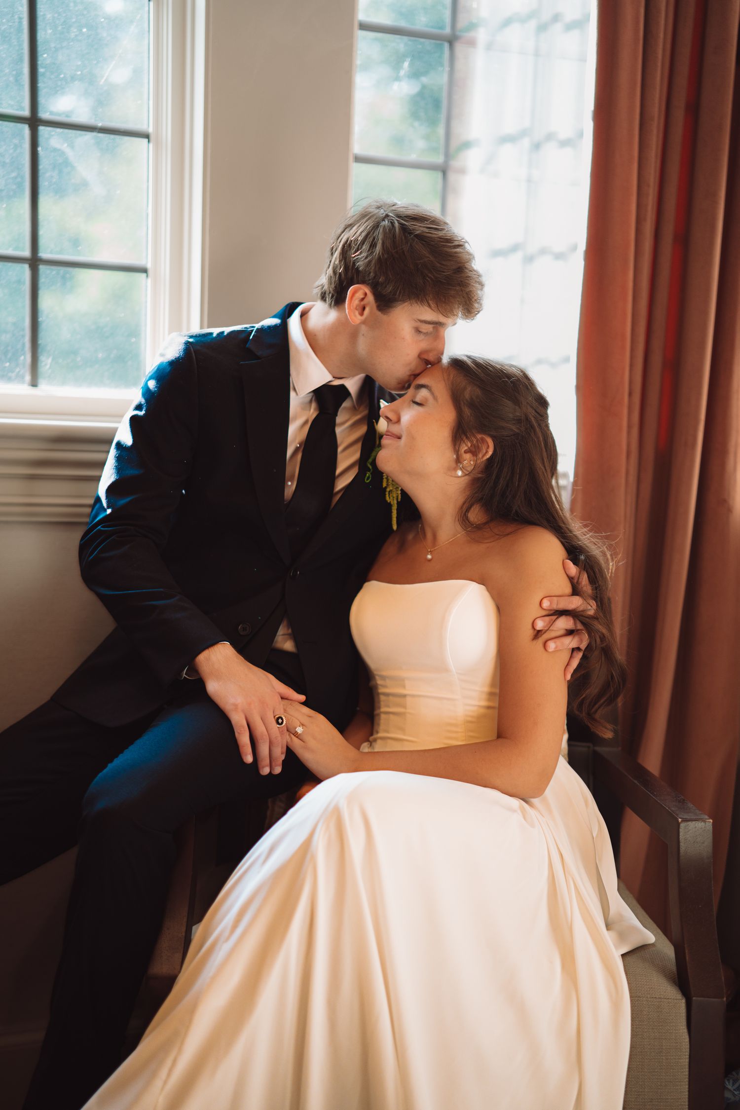 A groom in a dark suit kisses his bride in a white strapless gown near a sunlit window.