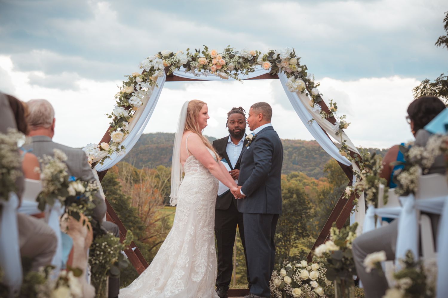 Outdoor wedding ceremony with bride and groom at floral arch against mountain backdrop.