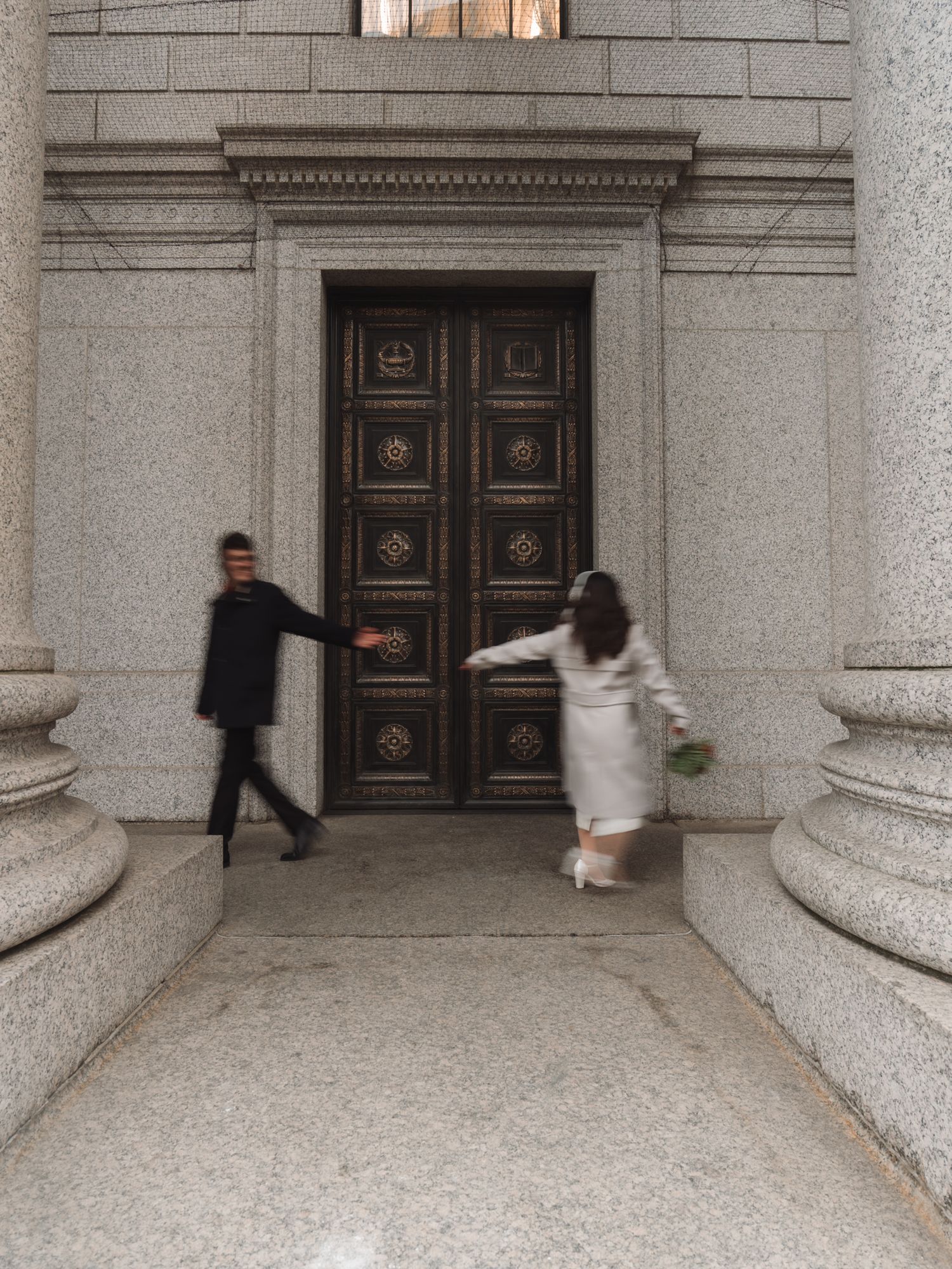 Newlyweds rushing through grand ornate bronze doors of a neoclassical building together.