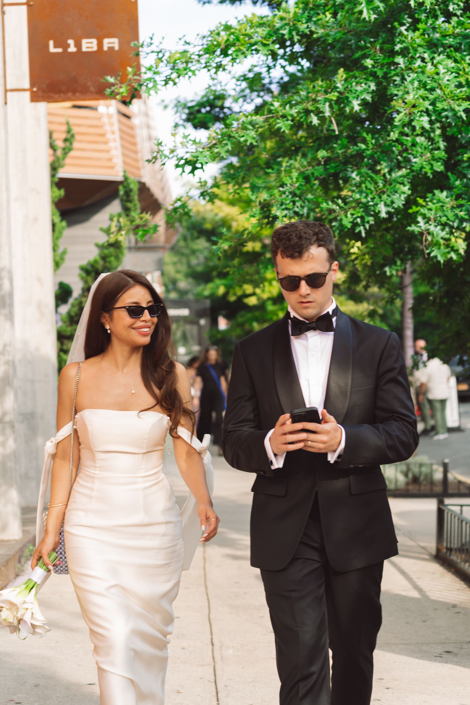 Bride in white gown and groom in black tuxedo walking outdoors, both wearing sunglasses, groom looking at phone.