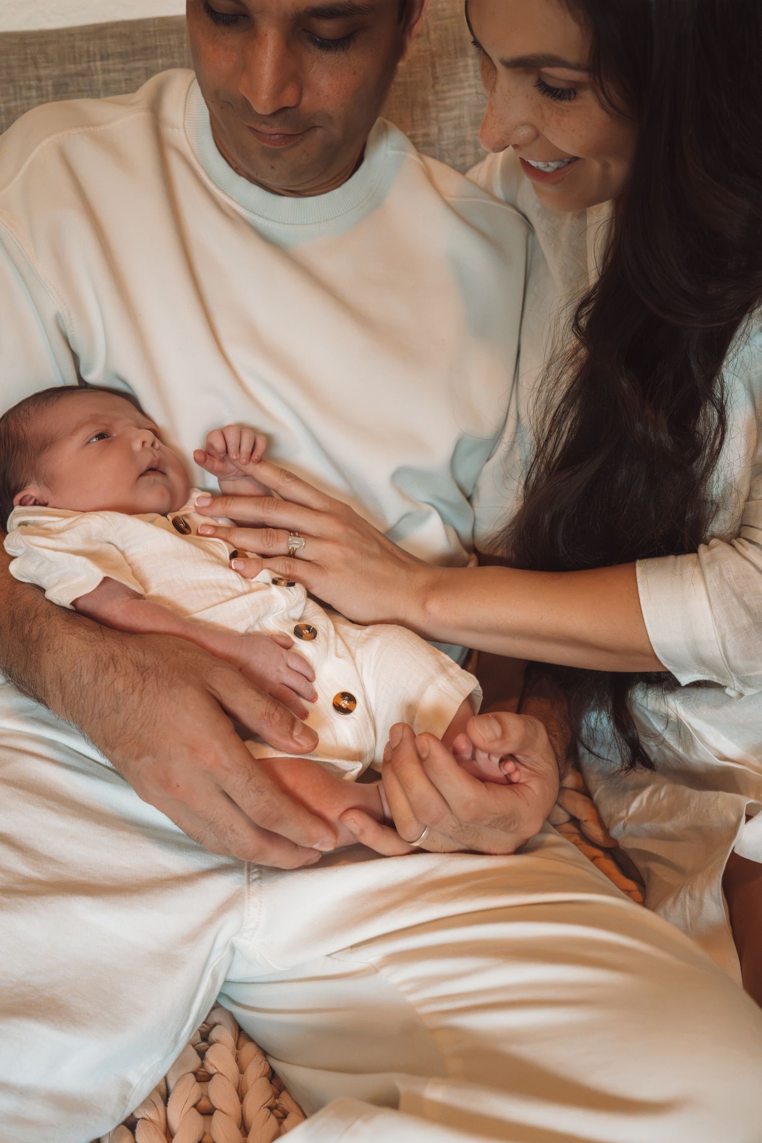 A couple tenderly holds their newborn baby together, sharing a joyful moment in soft, light-colored clothing.