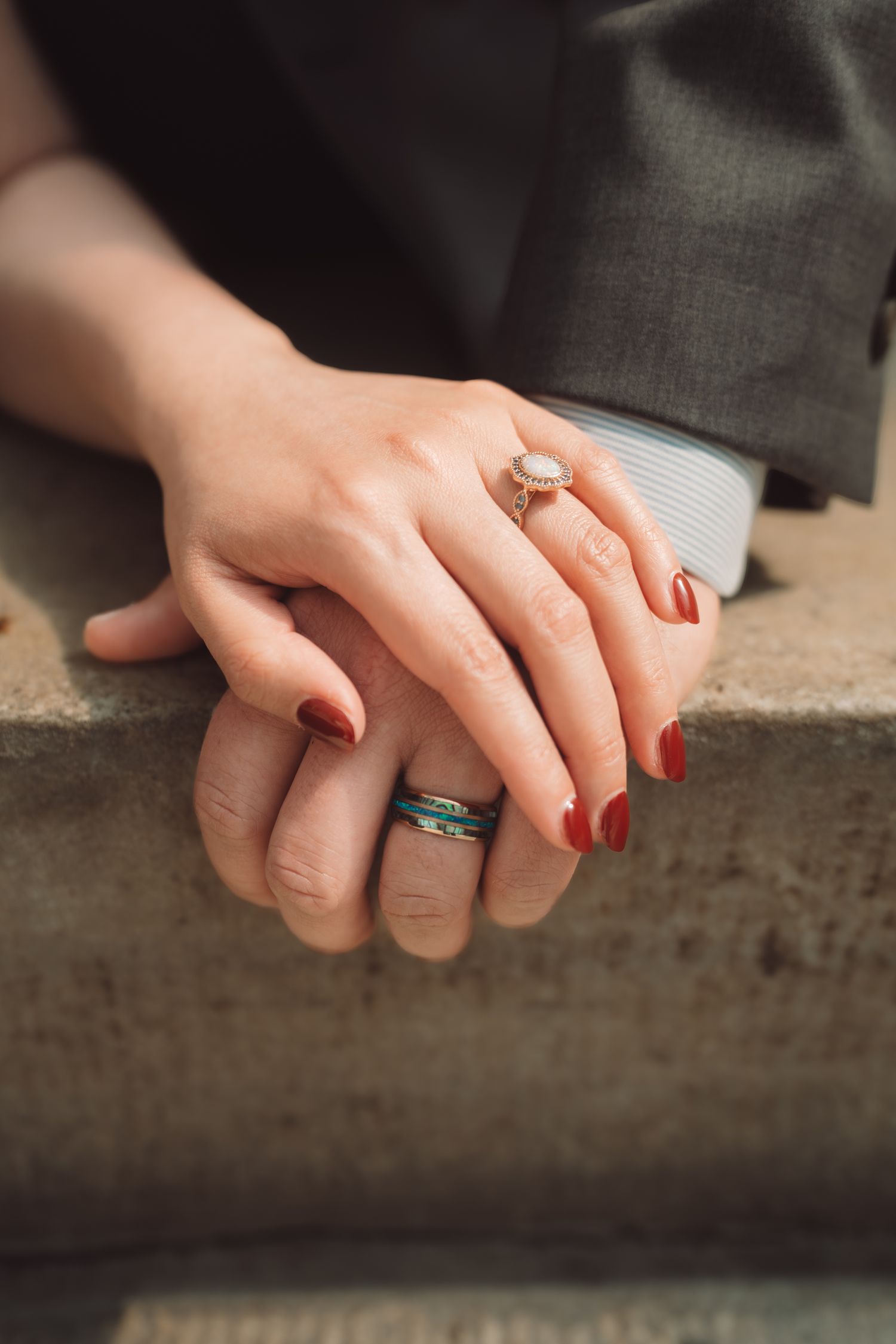 Close-up of couple's hands resting together, woman wearing pearl engagement ring with red nails, man wearing stacked rings.