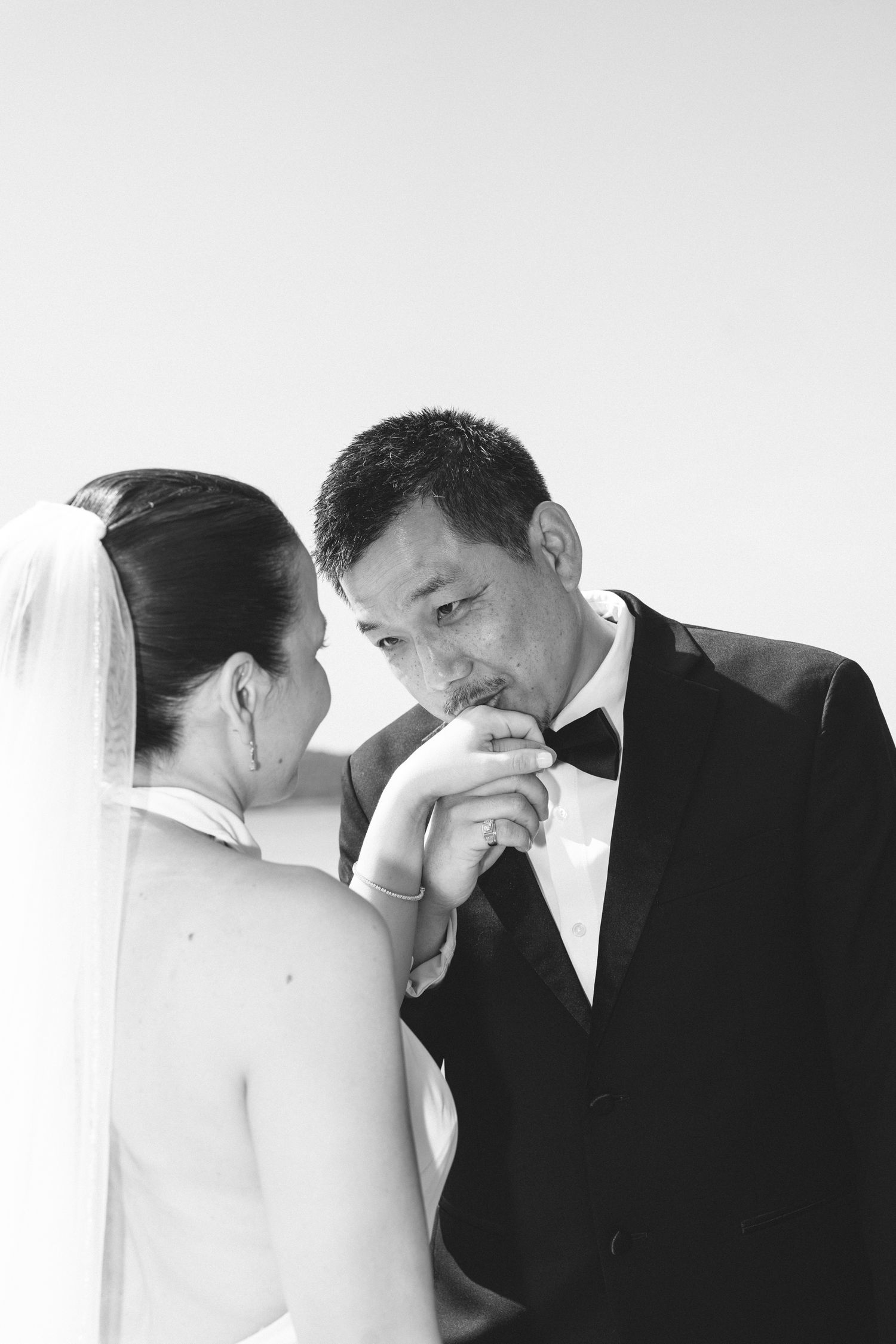 Groom in black tuxedo kissing bride's hand in black and white wedding photo.
