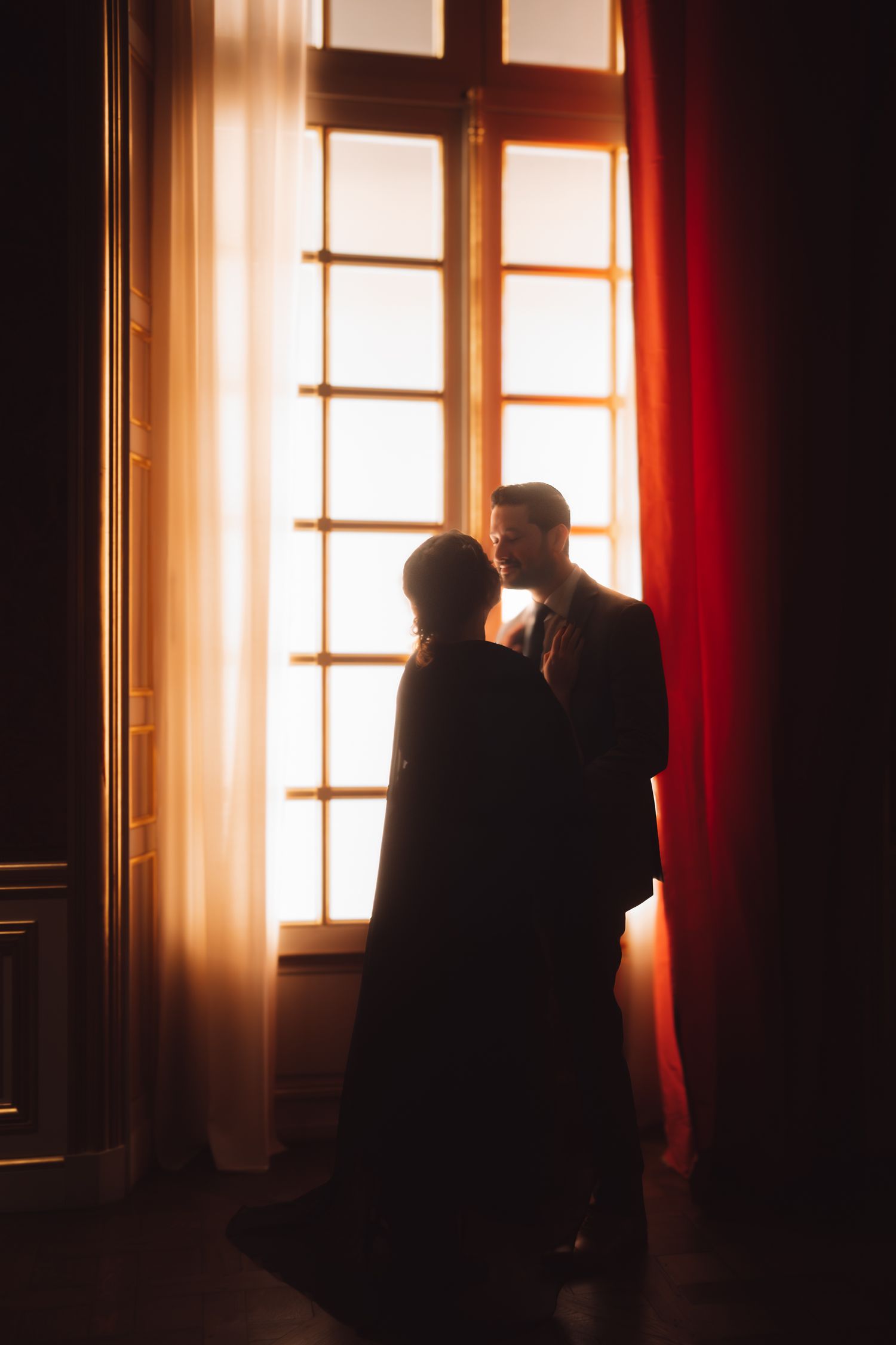 Romantic couple silhouetted against a glowing window with red curtains in an elegant room.