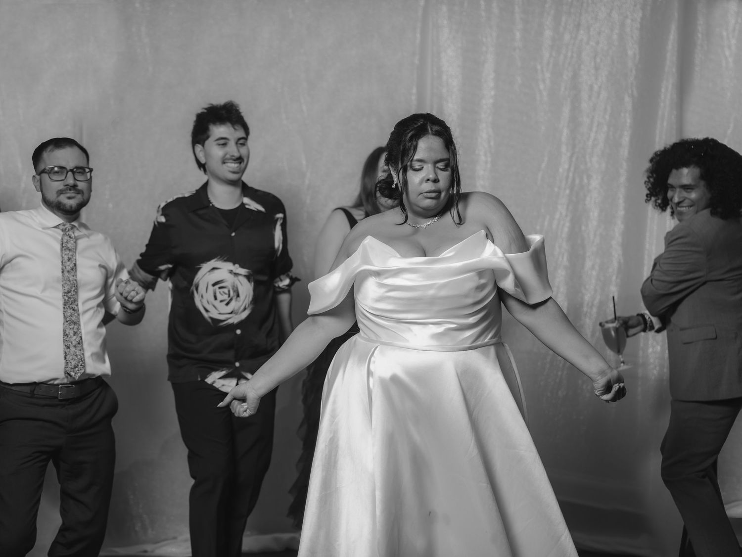 Bride in white satin gown dancing joyfully with guests at wedding reception in black and white photo.