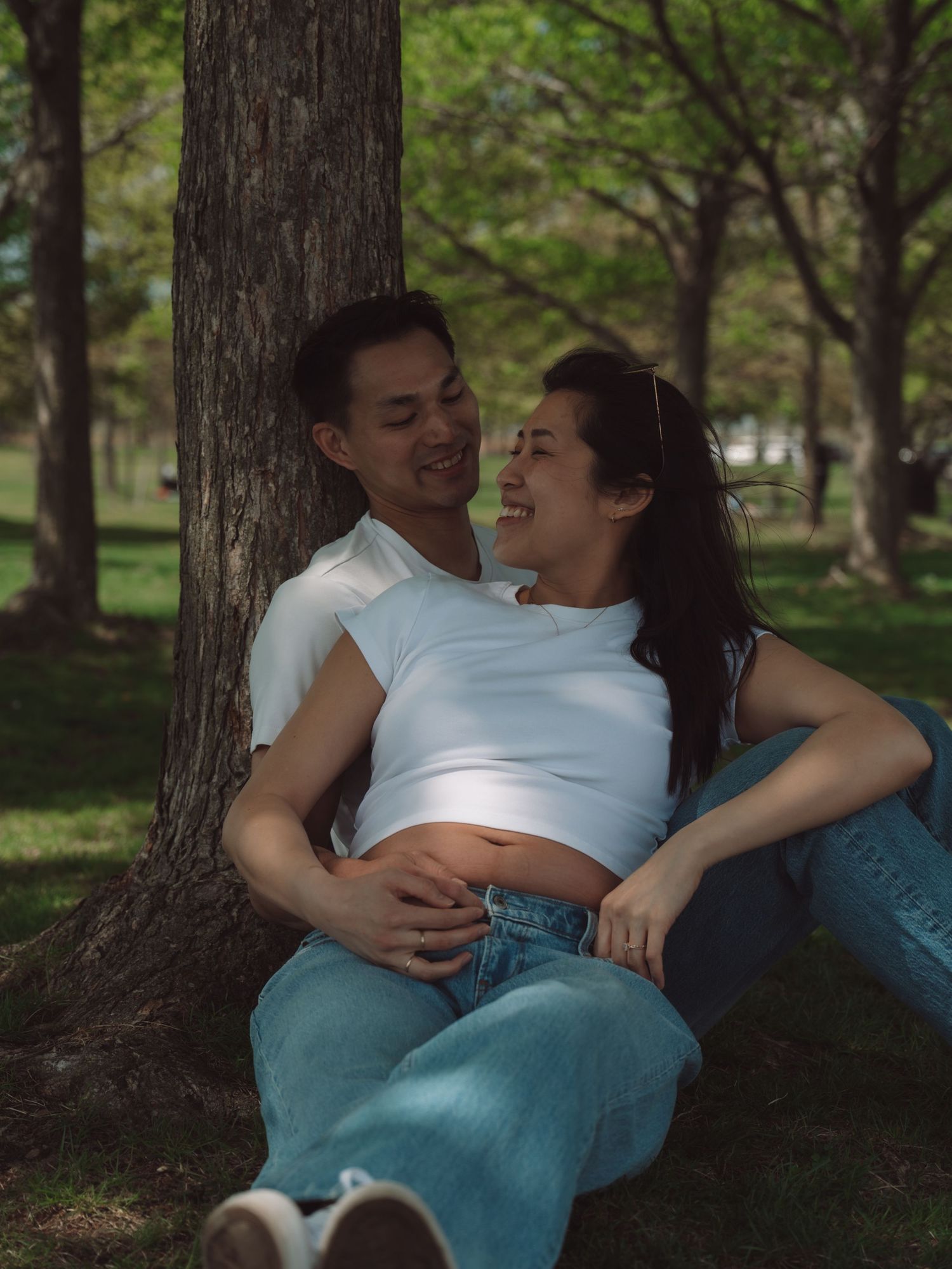 A couple sits together against a tree in a park, the woman showing a baby bump, both smiling joyfully.