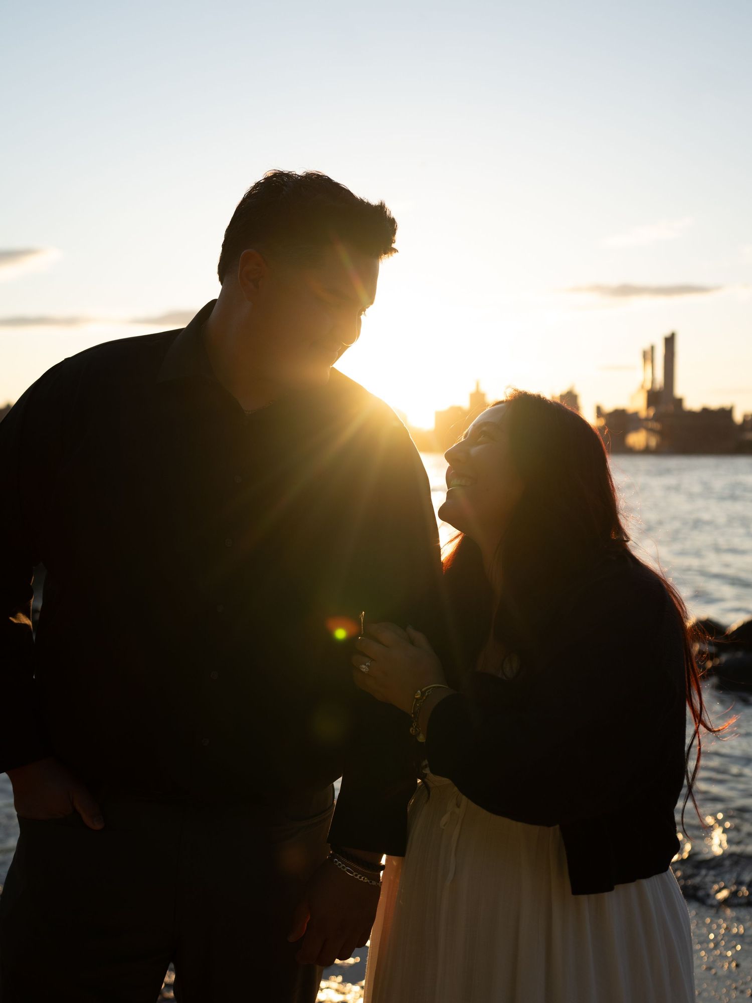 Romantic couple silhouetted against golden sunset by the waterfront, sharing an intimate moment with city skyline in background.
