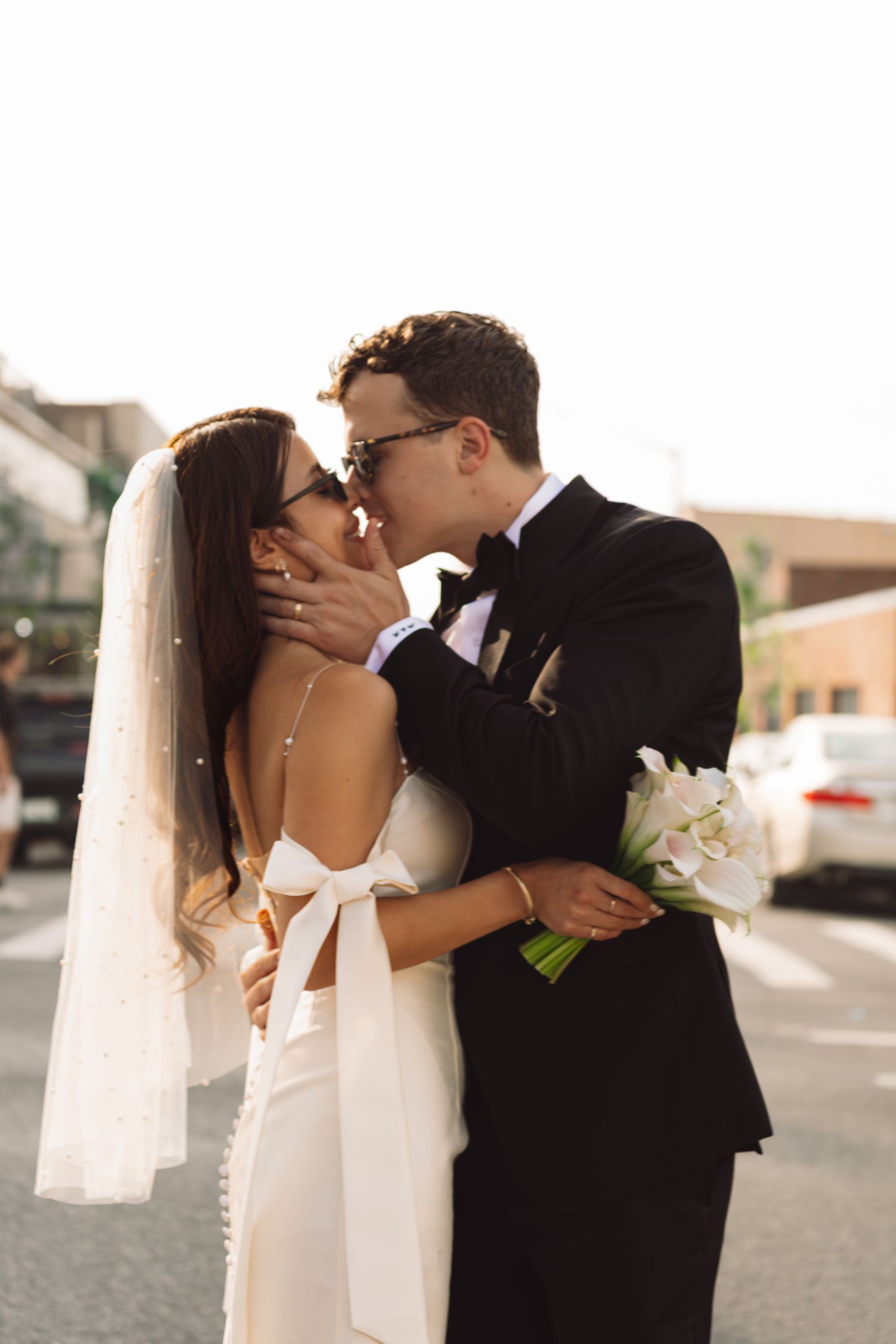 Bride and groom sharing a romantic kiss outdoors, bride in white gown with veil holding calla lily bouquet.