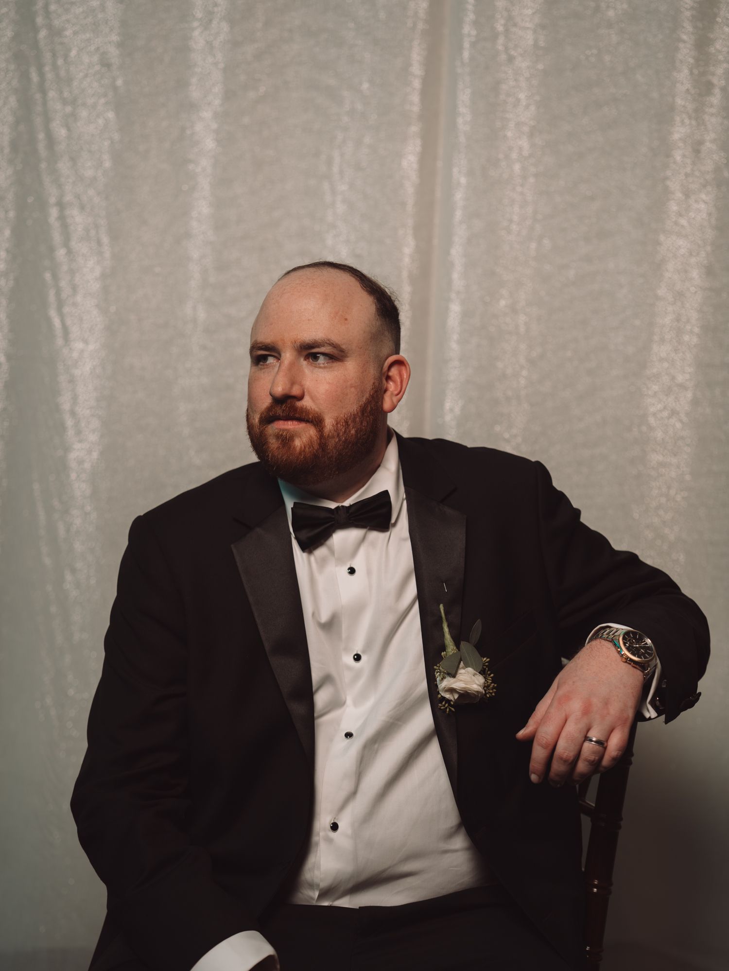Groom in black tuxedo with bow tie and boutonniere seated confidently at wedding reception.