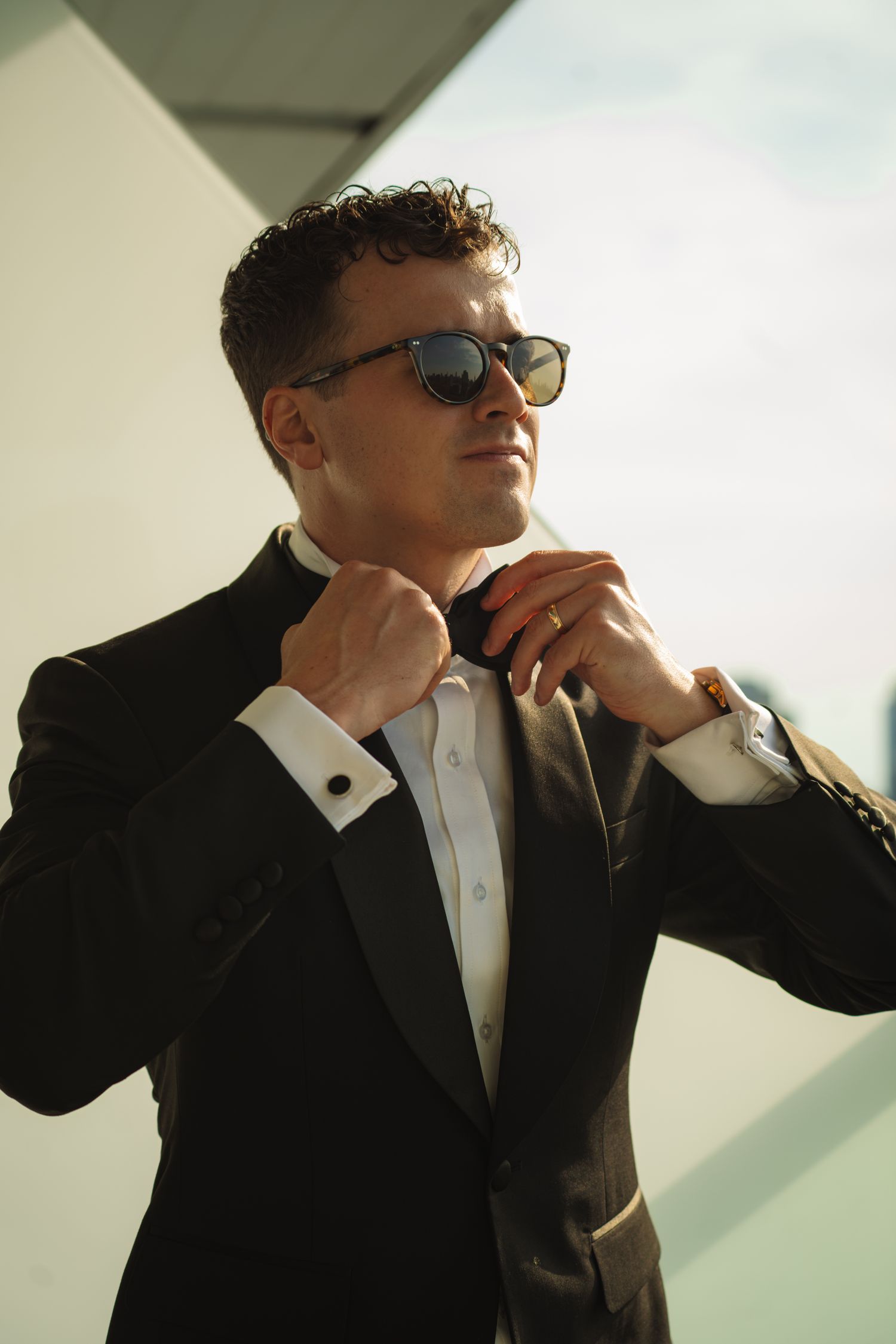 A stylish man in a black tuxedo and sunglasses adjusts his bow tie outdoors in bright sunlight.