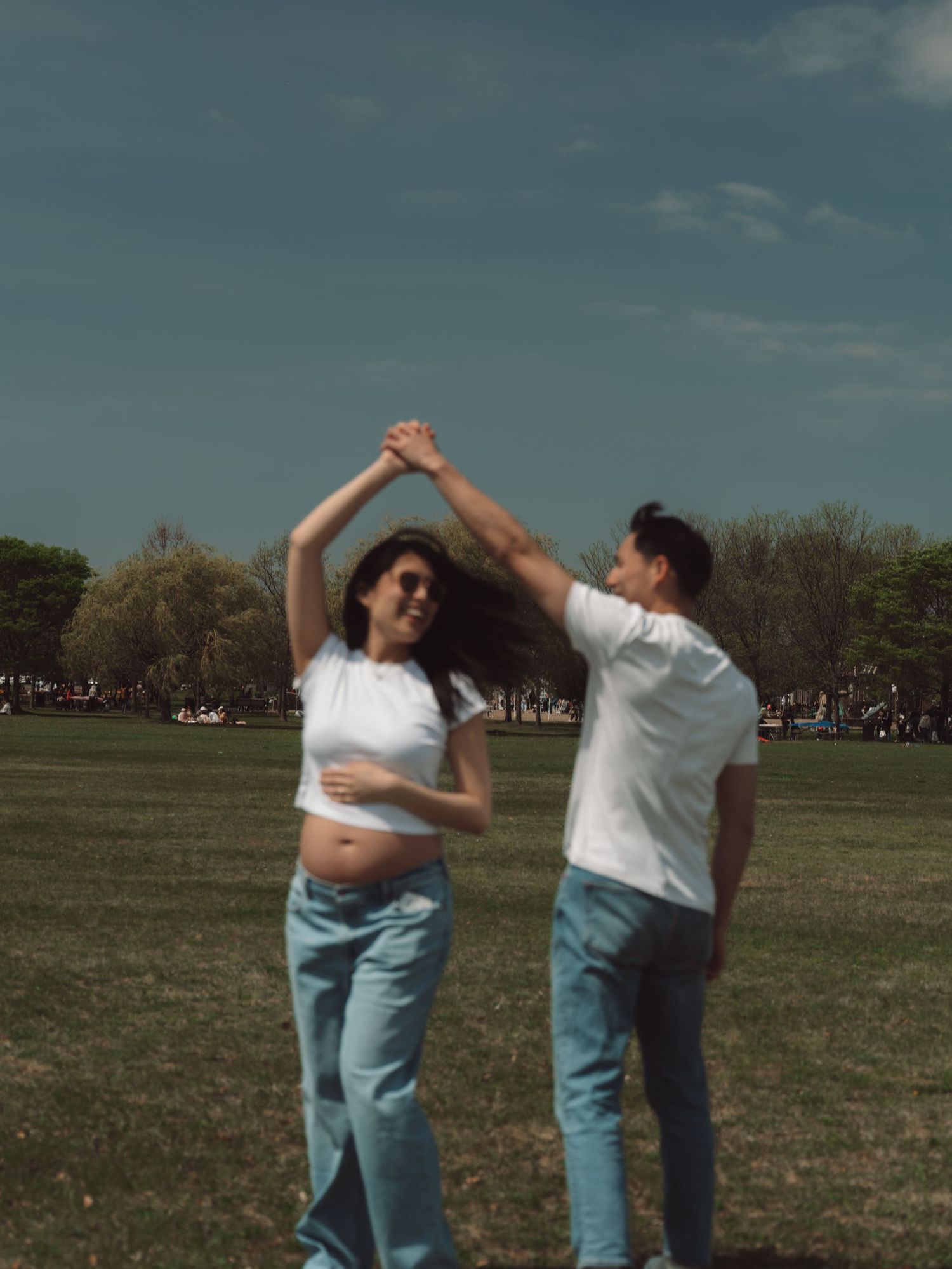 Pregnant couple dancing together in a sunny park, both wearing white shirts and light blue jeans.