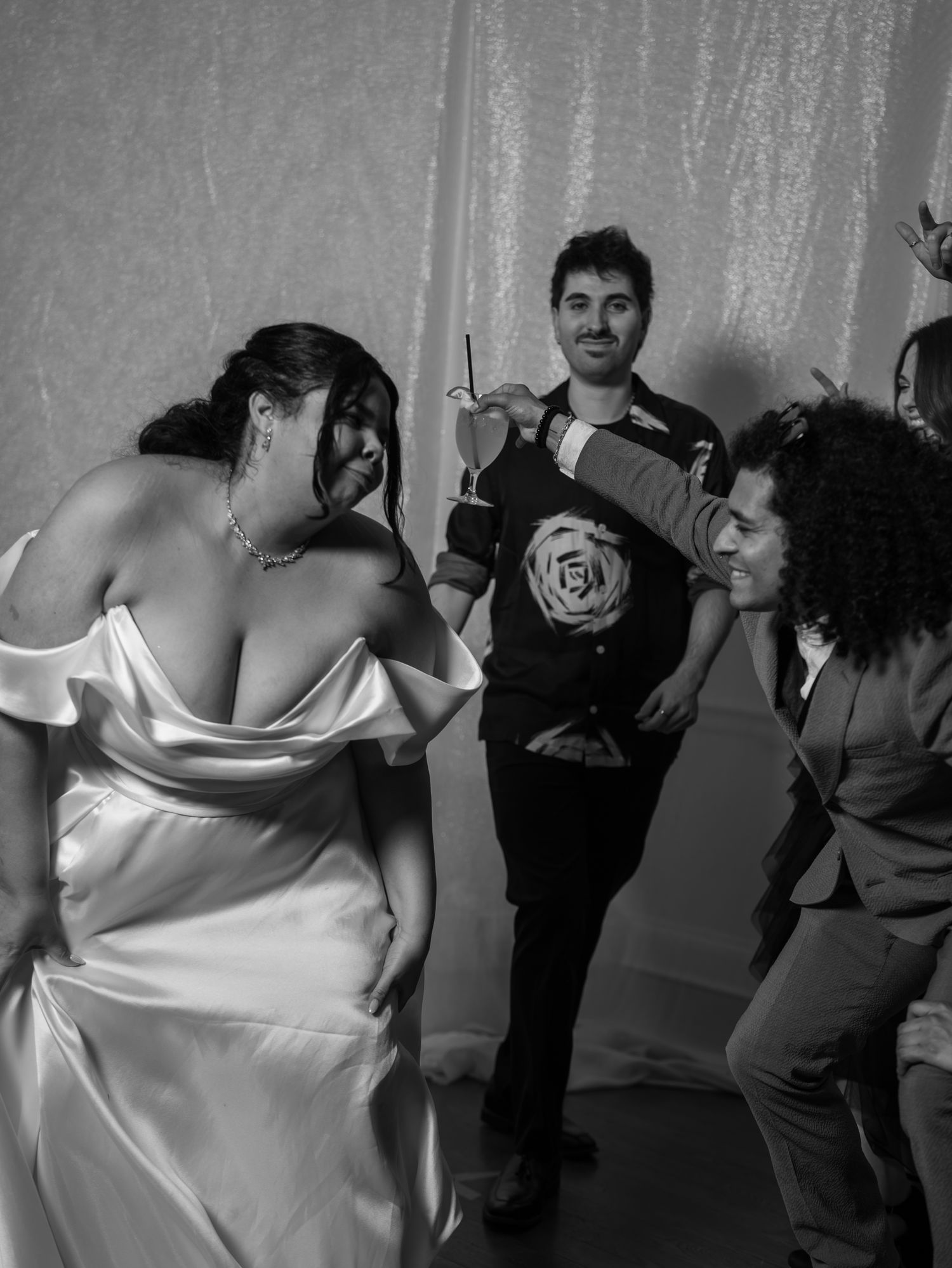 Black and white photo of bride in white satin gown dancing with guests at wedding reception.