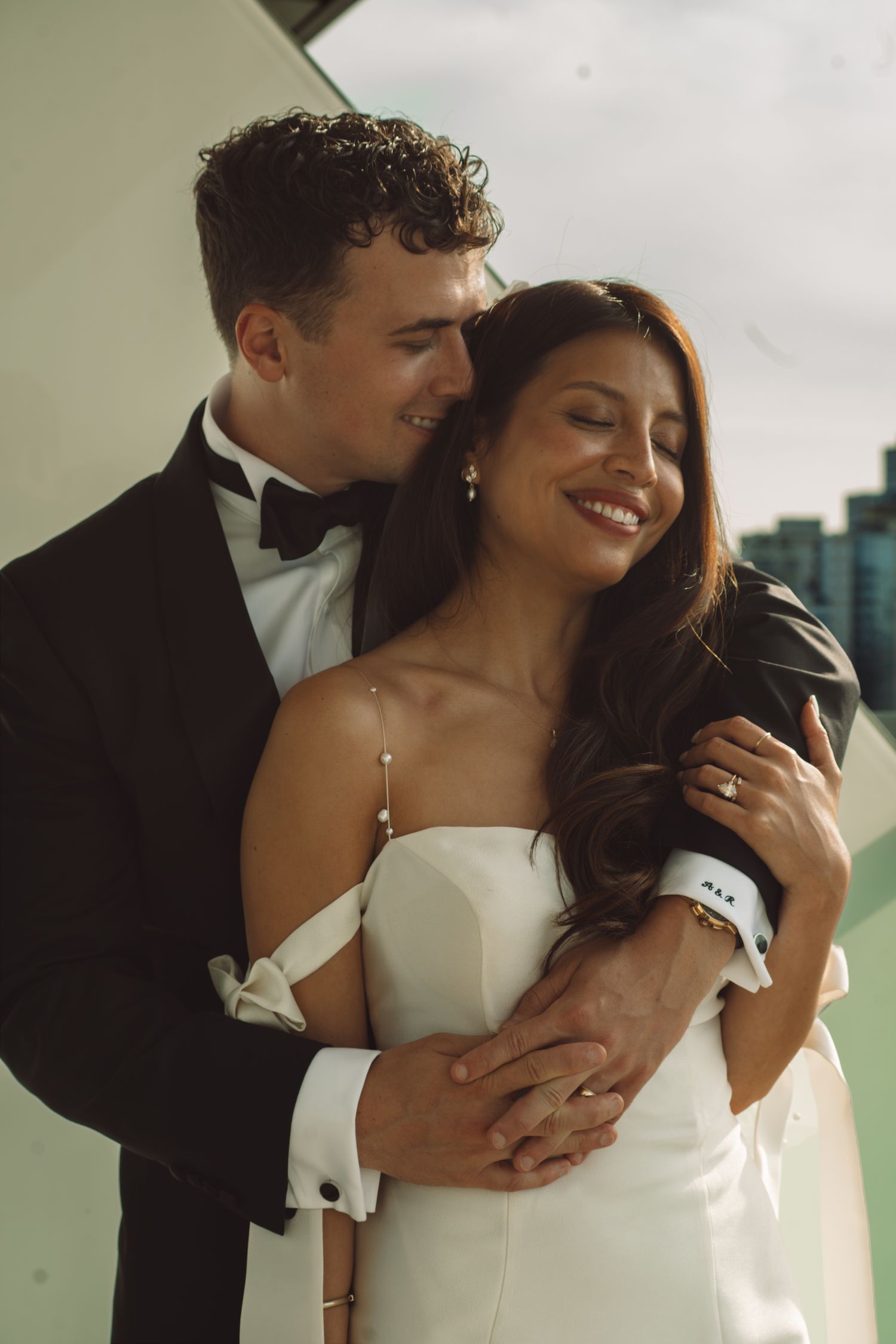A groom in a black tuxedo embraces his smiling bride in a white off-shoulder gown during their wedding day.