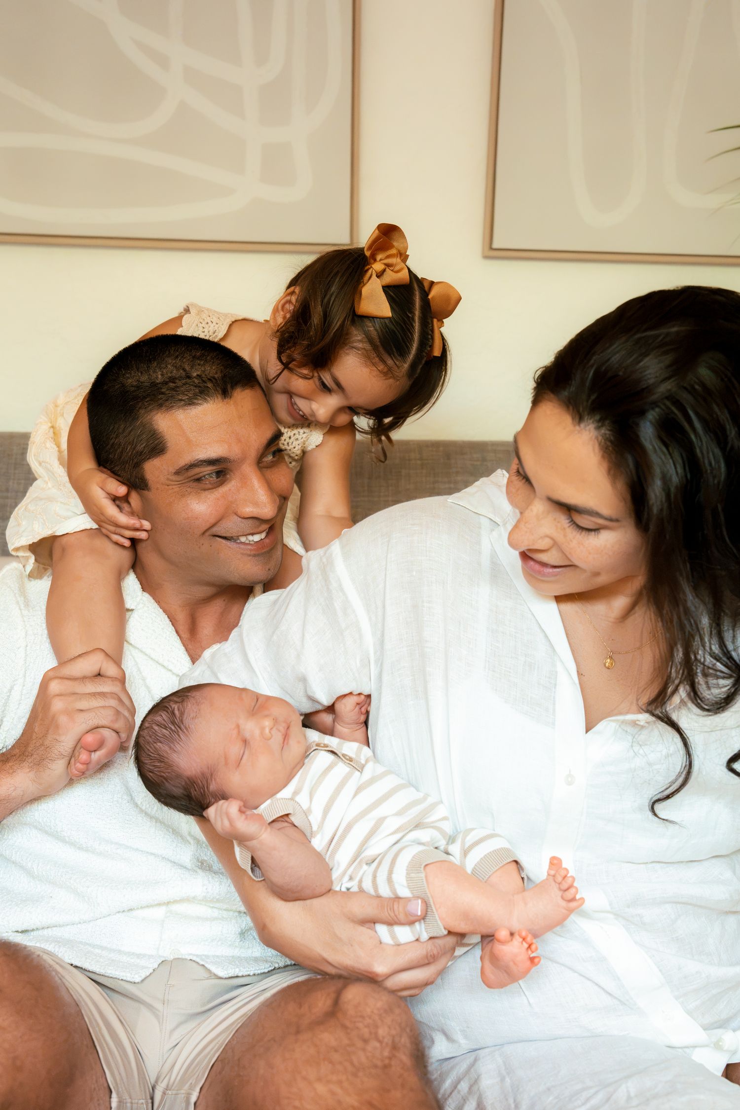 Happy family with parents in white holding newborn baby while toddler girl with bow kisses dad on cheek.