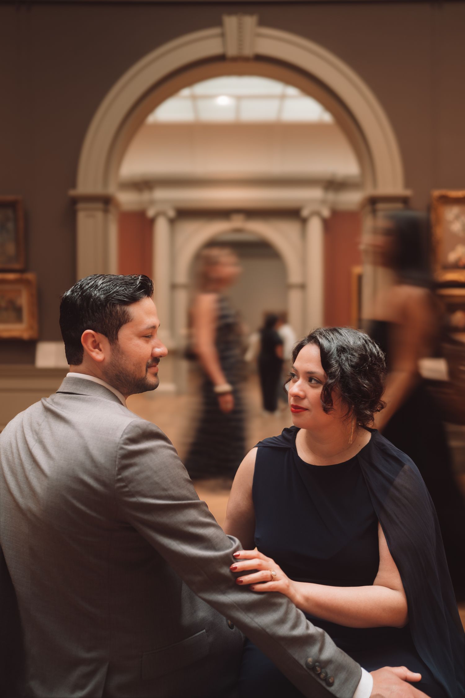 Couple sharing an intimate moment at an elegant art museum gala, woman in navy dress, man in gray suit.